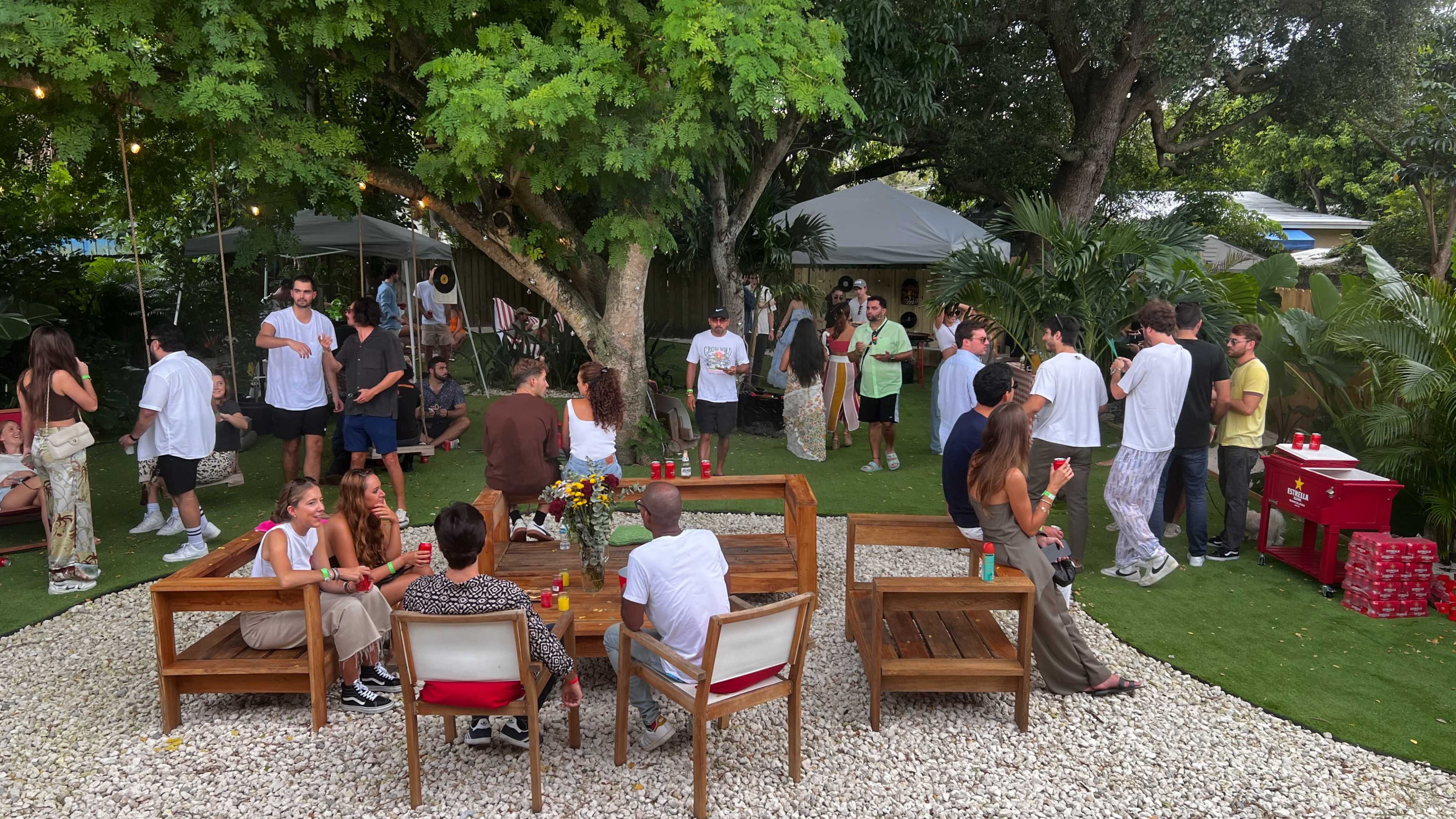 A garden party scene features groups of people socializing around wooden furniture, surrounded by lush greenery and decorative elements.