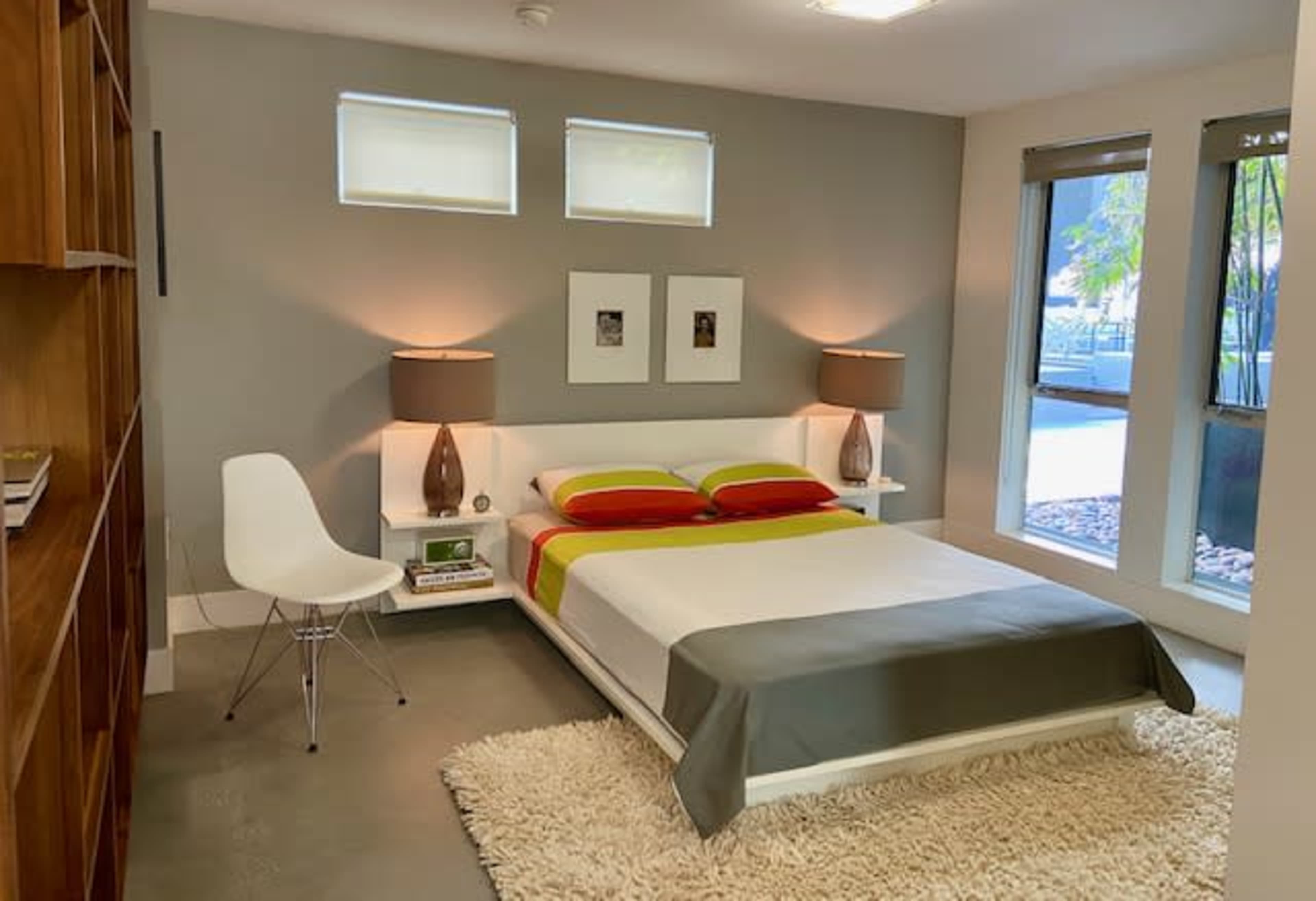A modern bedroom featuring a white bed with colorful pillows, two lamps on either side, and large windows with a view.