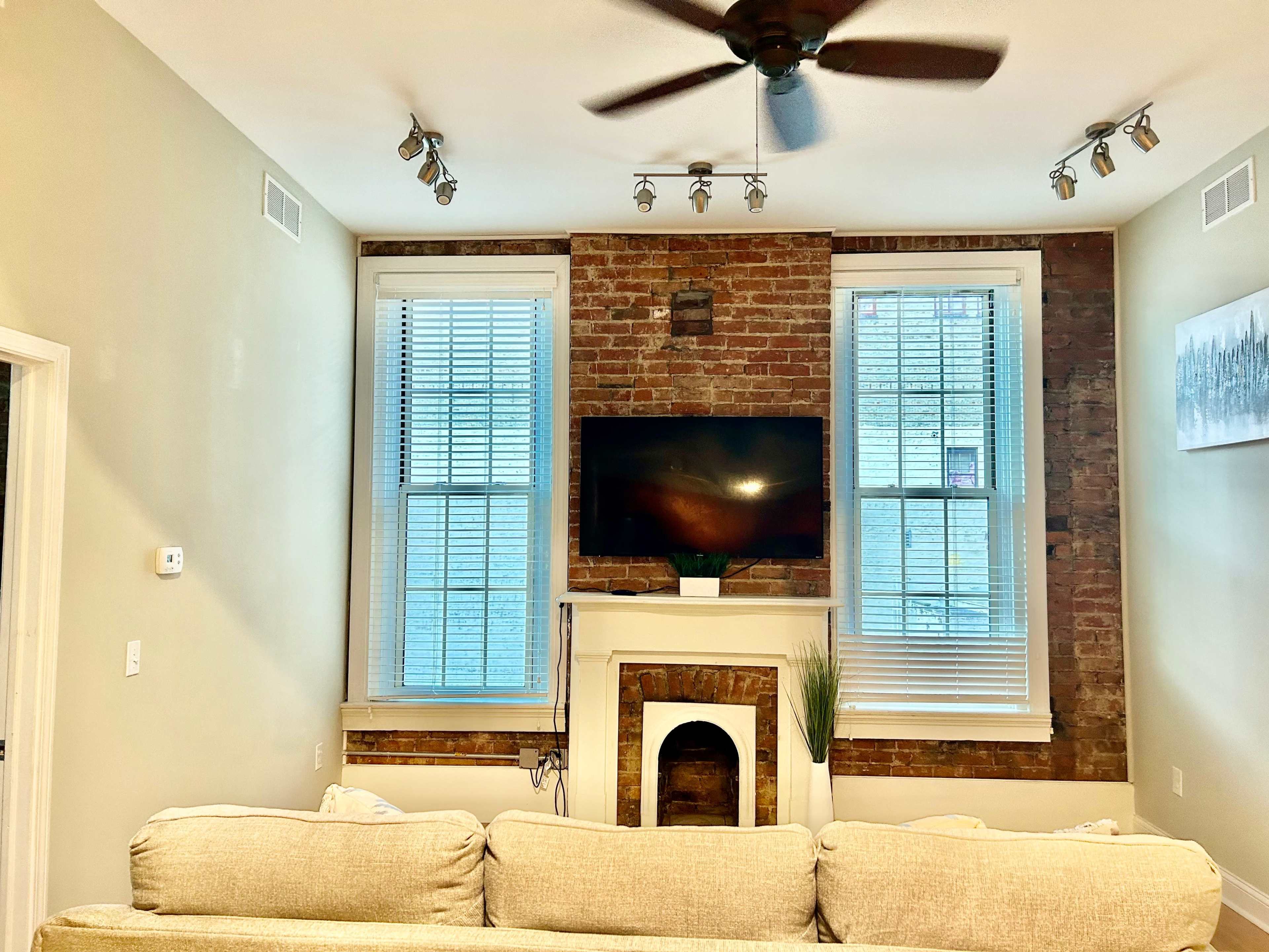 A living room with a brick accent wall, two large windows, a mounted television above a fireplace, and a beige couch in the foreground.