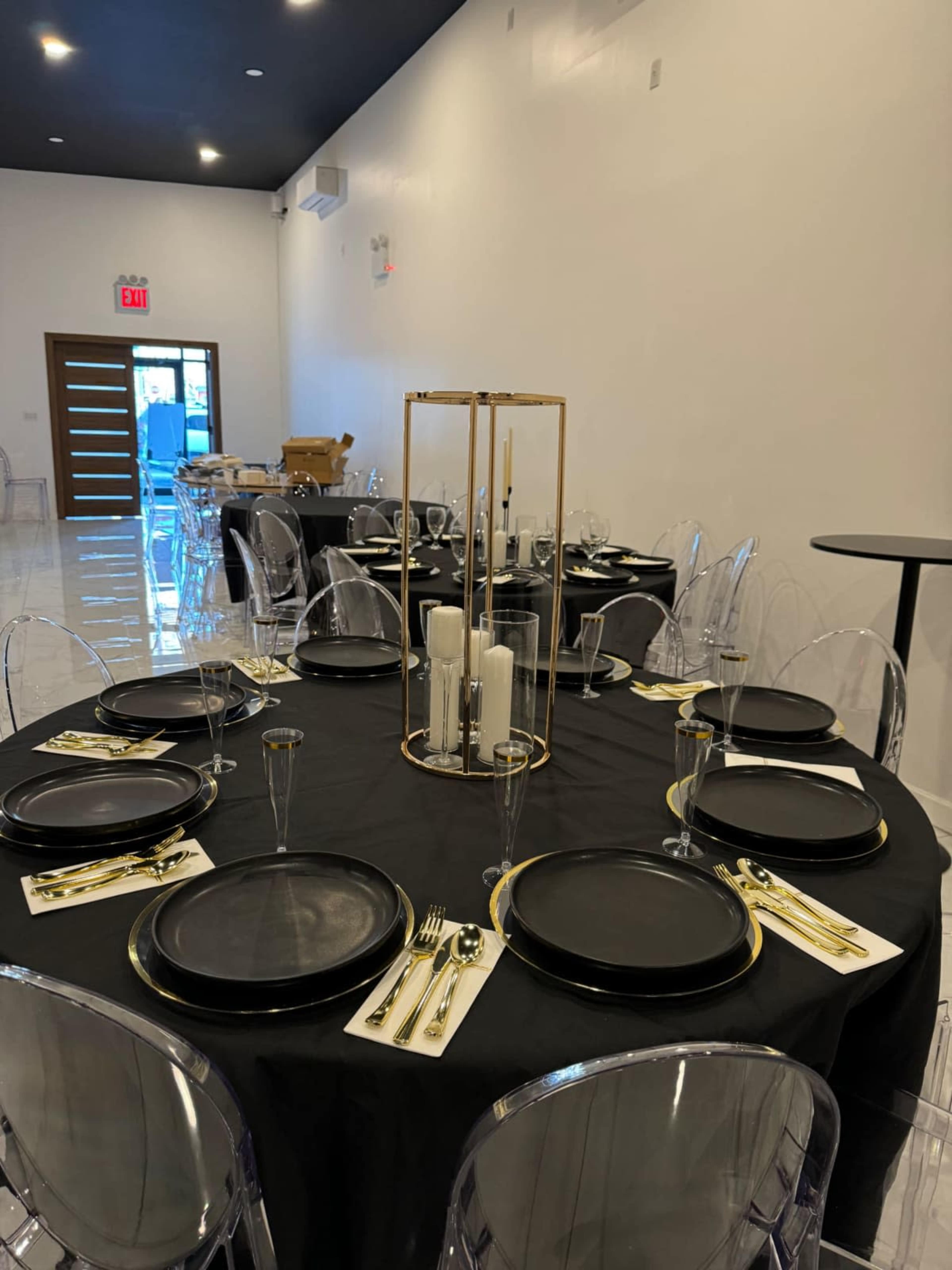 A dining area is set with black tablecloths, clear chairs, and tables arranged with circular plates and gold utensils.