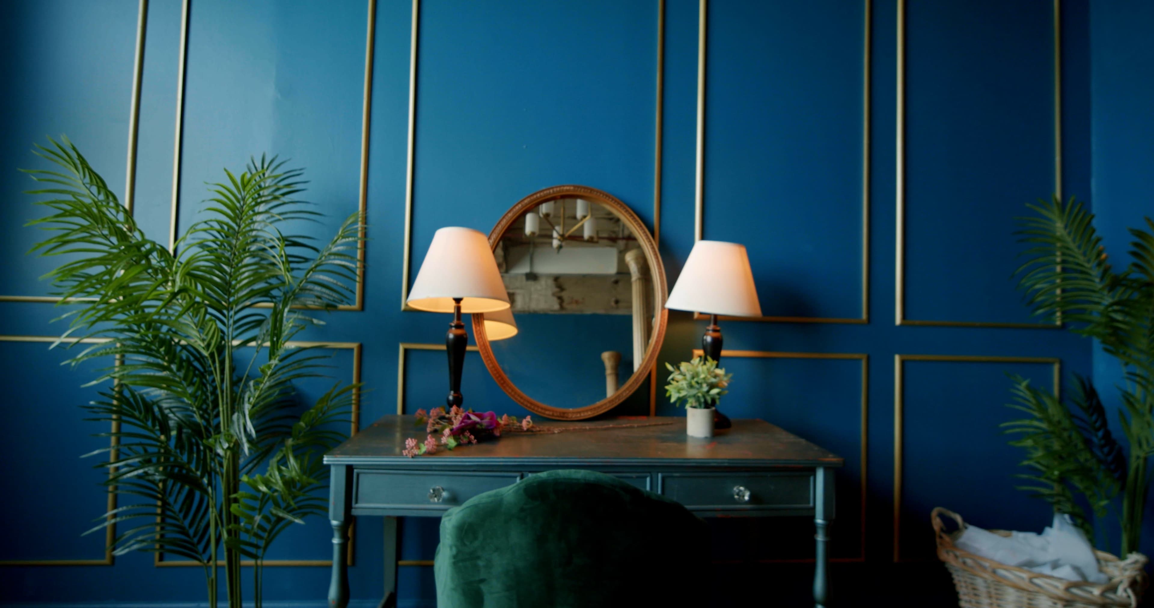 A green velvet chair sits at a wooden desk with a round mirror and two lamps against a blue wall adorned with gold paneling, complemented by potted plants.