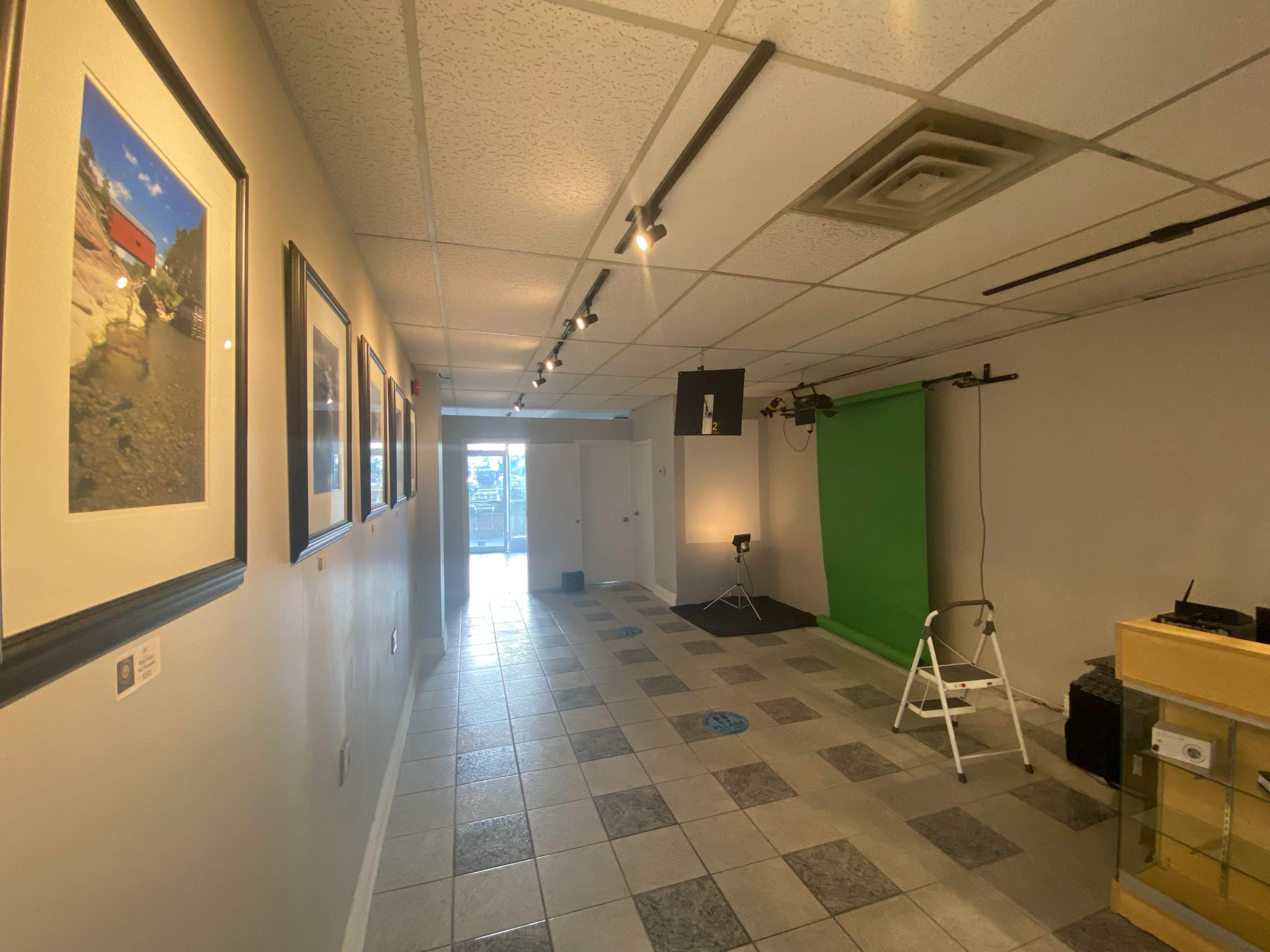 A hallway with framed photographs on the left, a green backdrop for video or photography at the far end, and a lighting setup on the right.