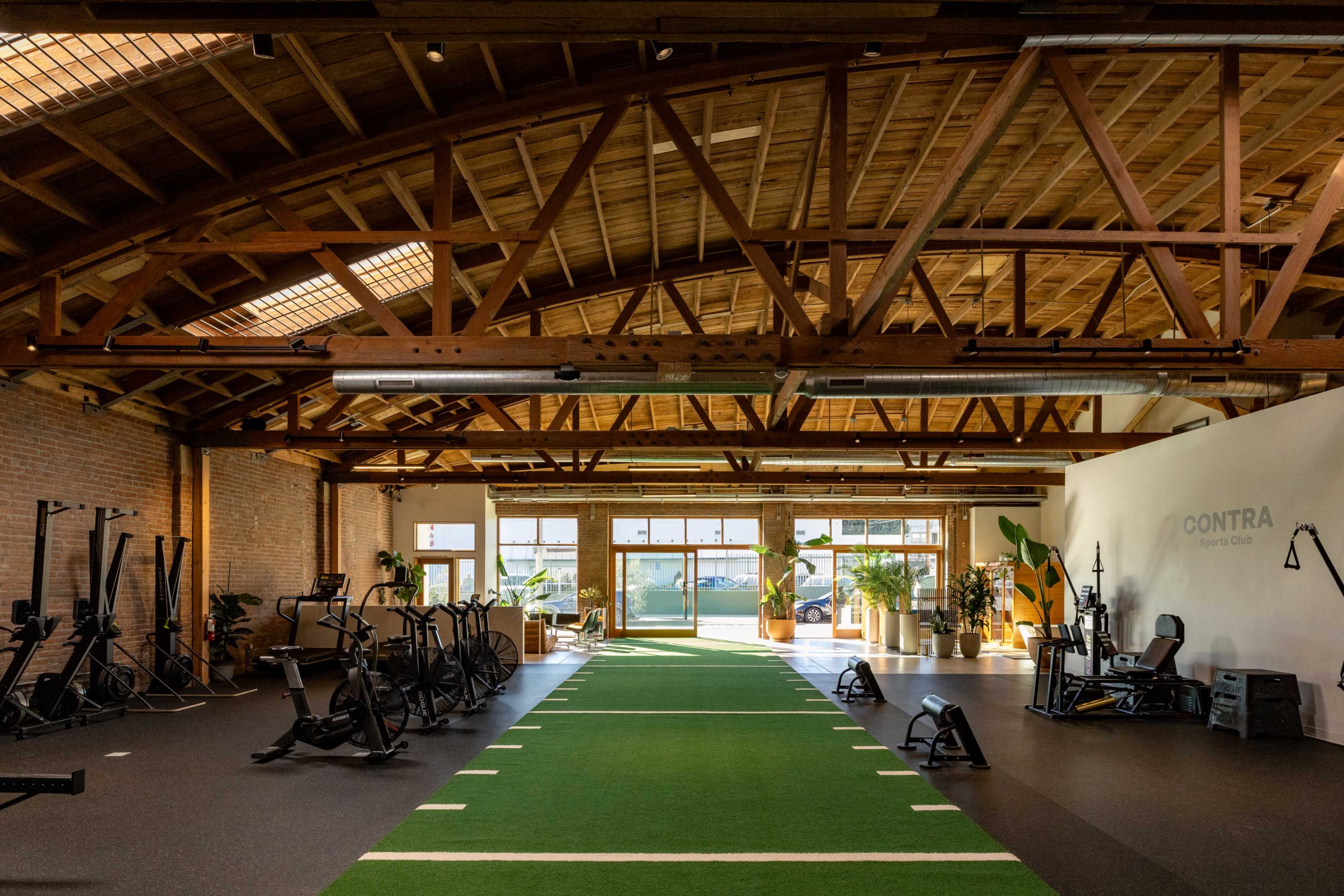 The image shows a spacious gym with wooden beams, equipped with fitness machines and a green turf area leading to large windows that allow natural light to fill the space.