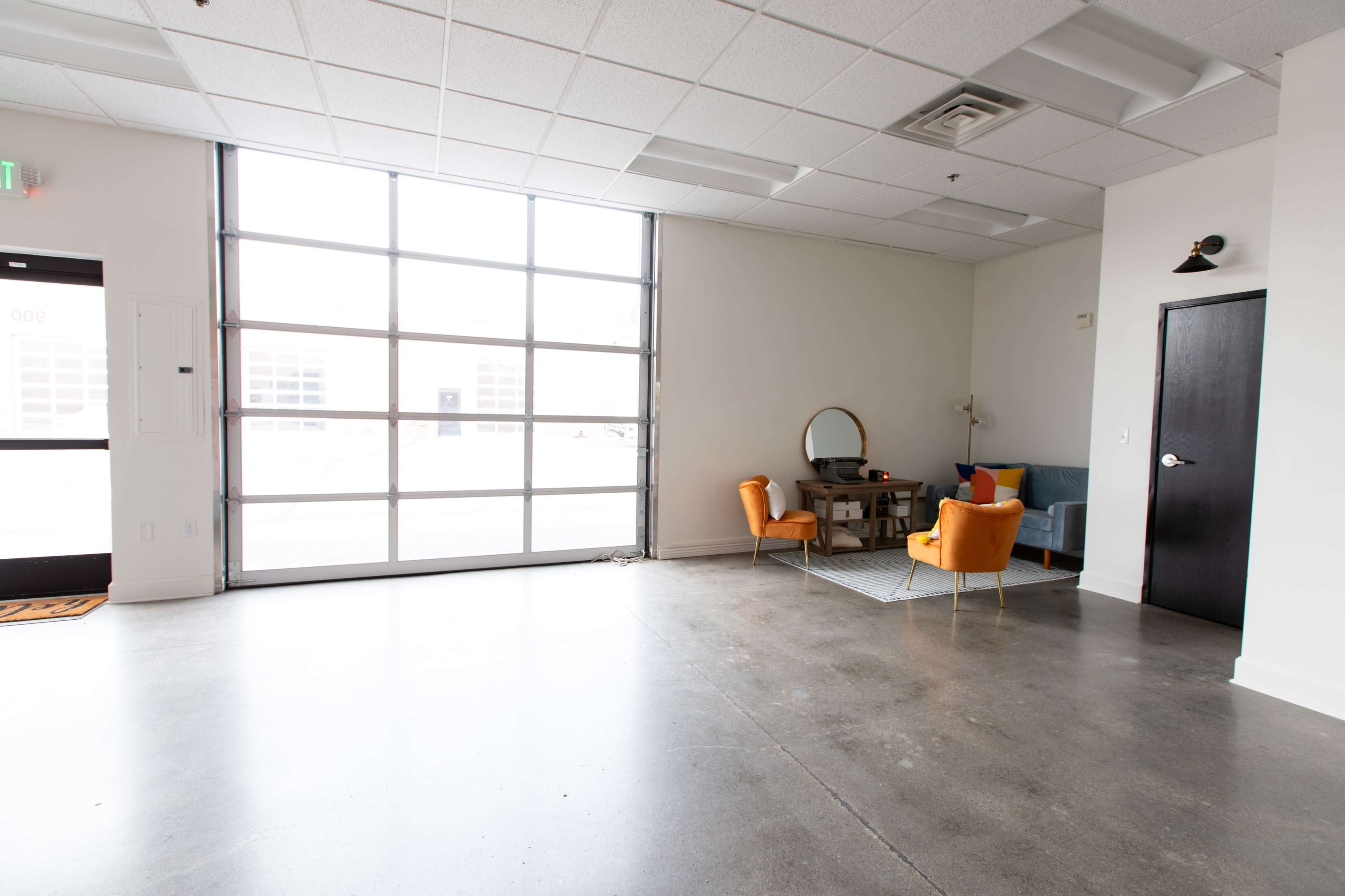 A spacious, empty room with a large window and two armchairs, a small table, and a mirror against the wall.
