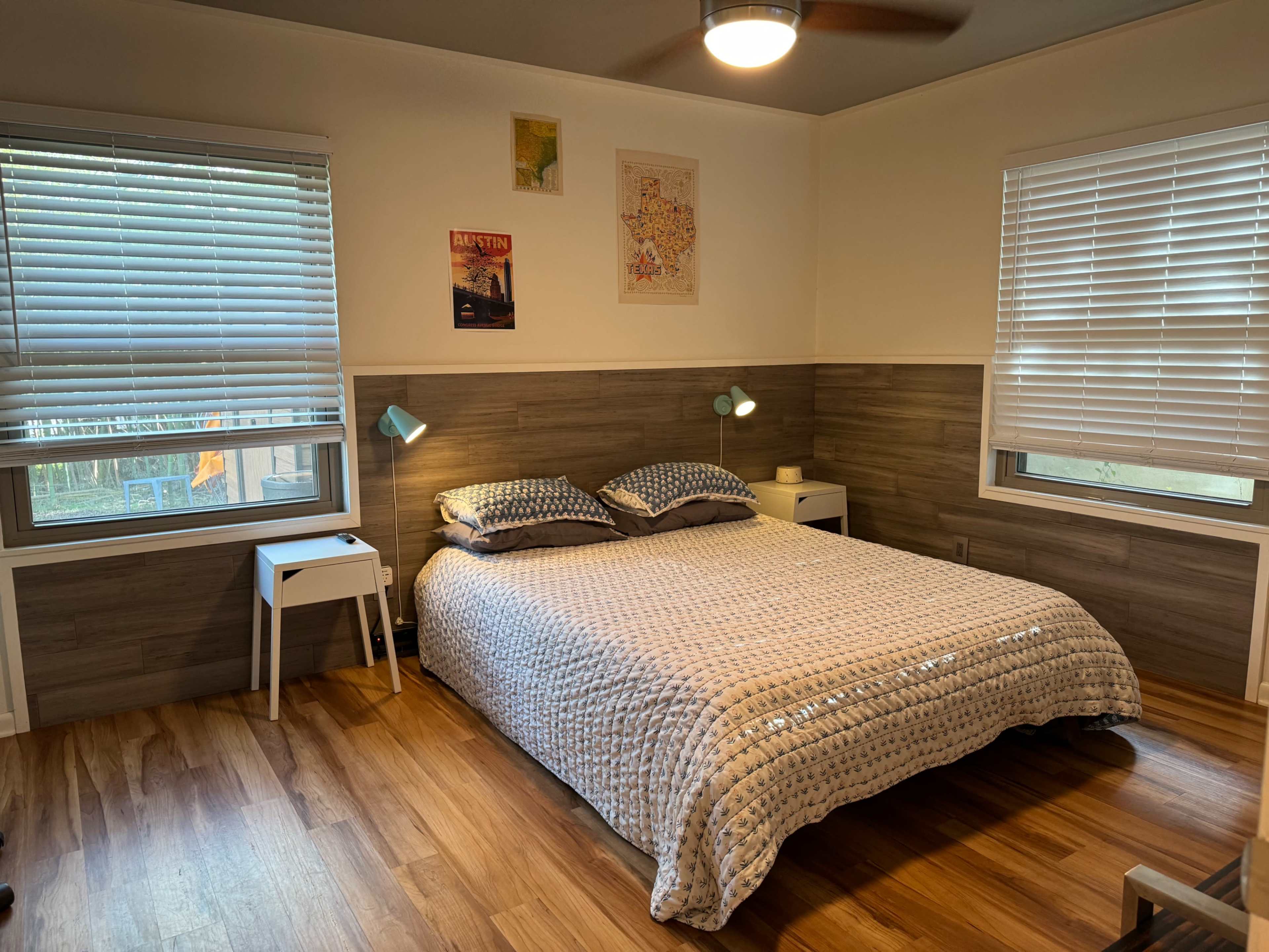 A neatly arranged bedroom featuring a double bed with a patterned quilt, two small nightstands with lamps, and windows with closed blinds.