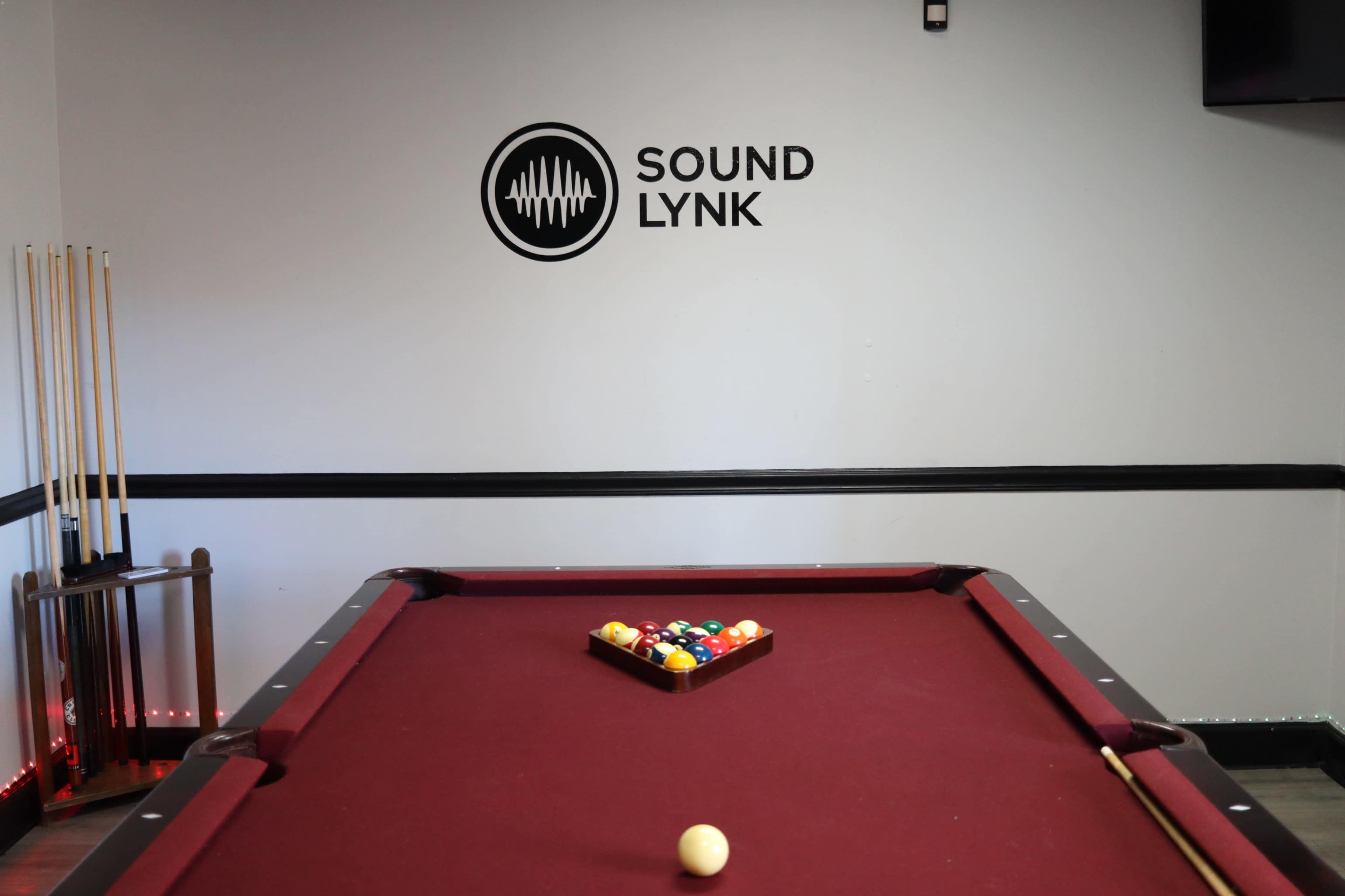 The image shows a red pool table with a triangle of colored balls arranged on it, set against a wall featuring the logo "SOUND LYNK."