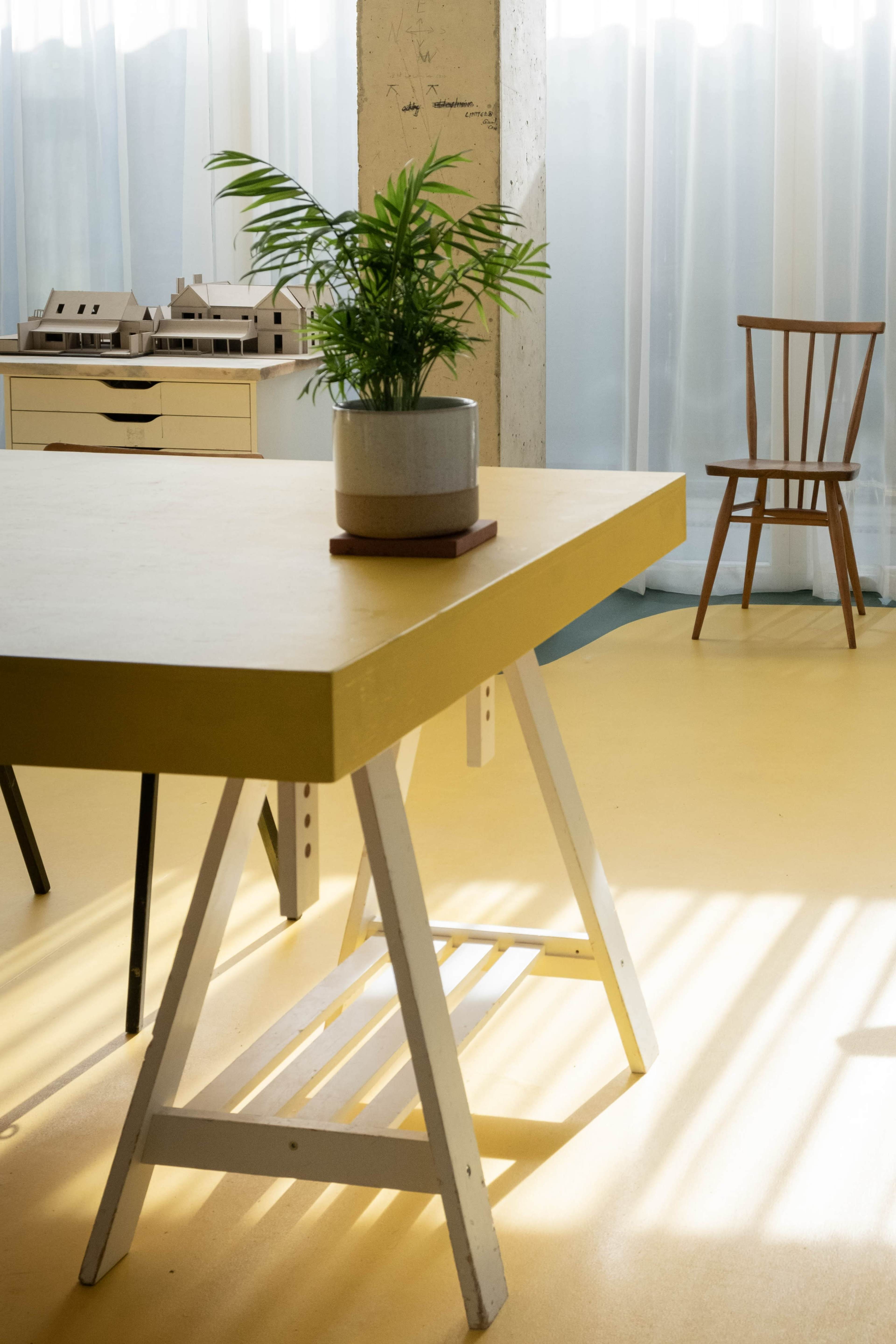 A yellow table with a potted plant sits on a light-colored floor beside a wooden chair and model houses.