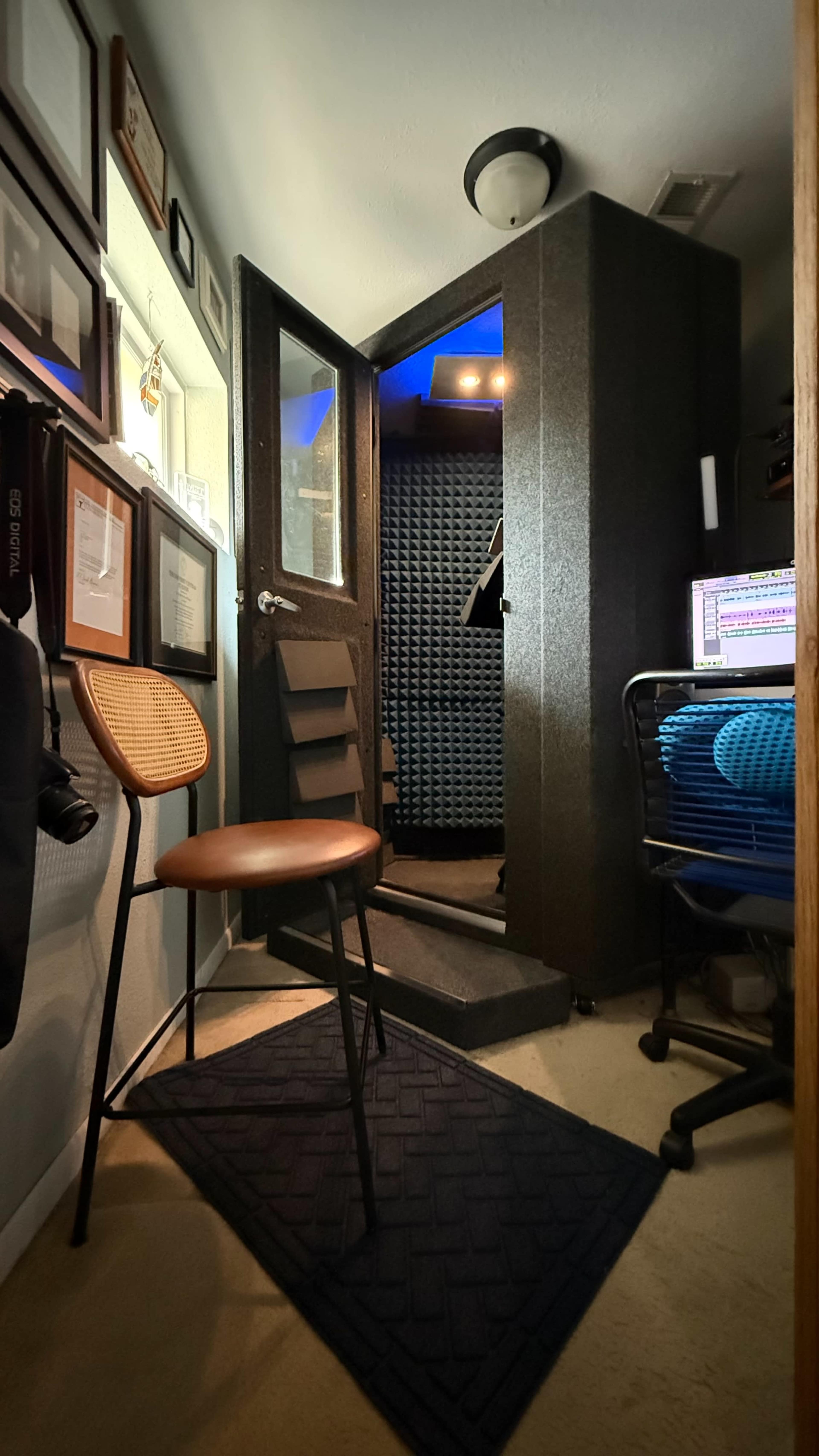 The image shows a small home recording studio with a soundproof booth, a chair, and a computer workstation.