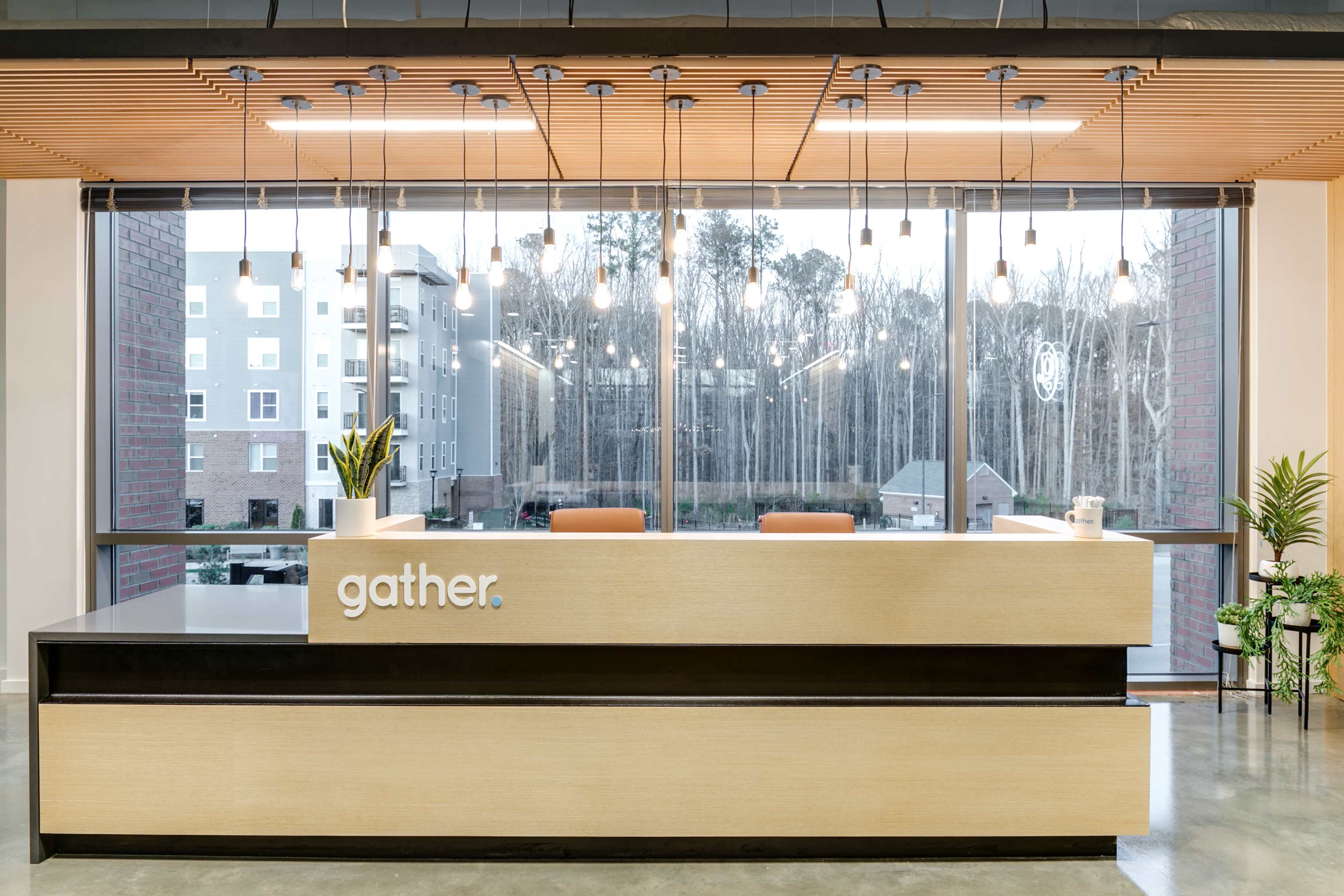 A modern reception desk with the word "gather" prominently displayed, illuminated by hanging light fixtures and large windows overlooking a wooded area.