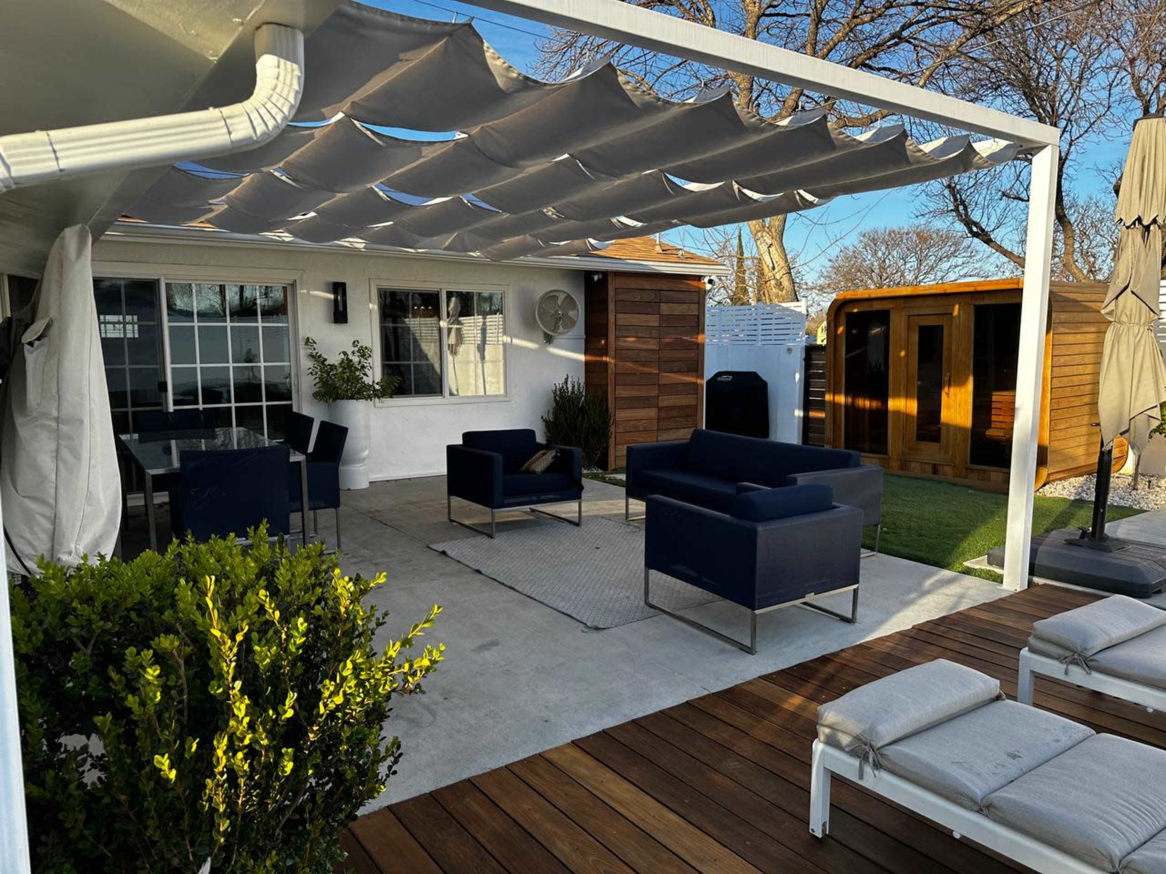 A modern outdoor patio area with a seating arrangement under a canopy, surrounded by greenery and small wooden structures.