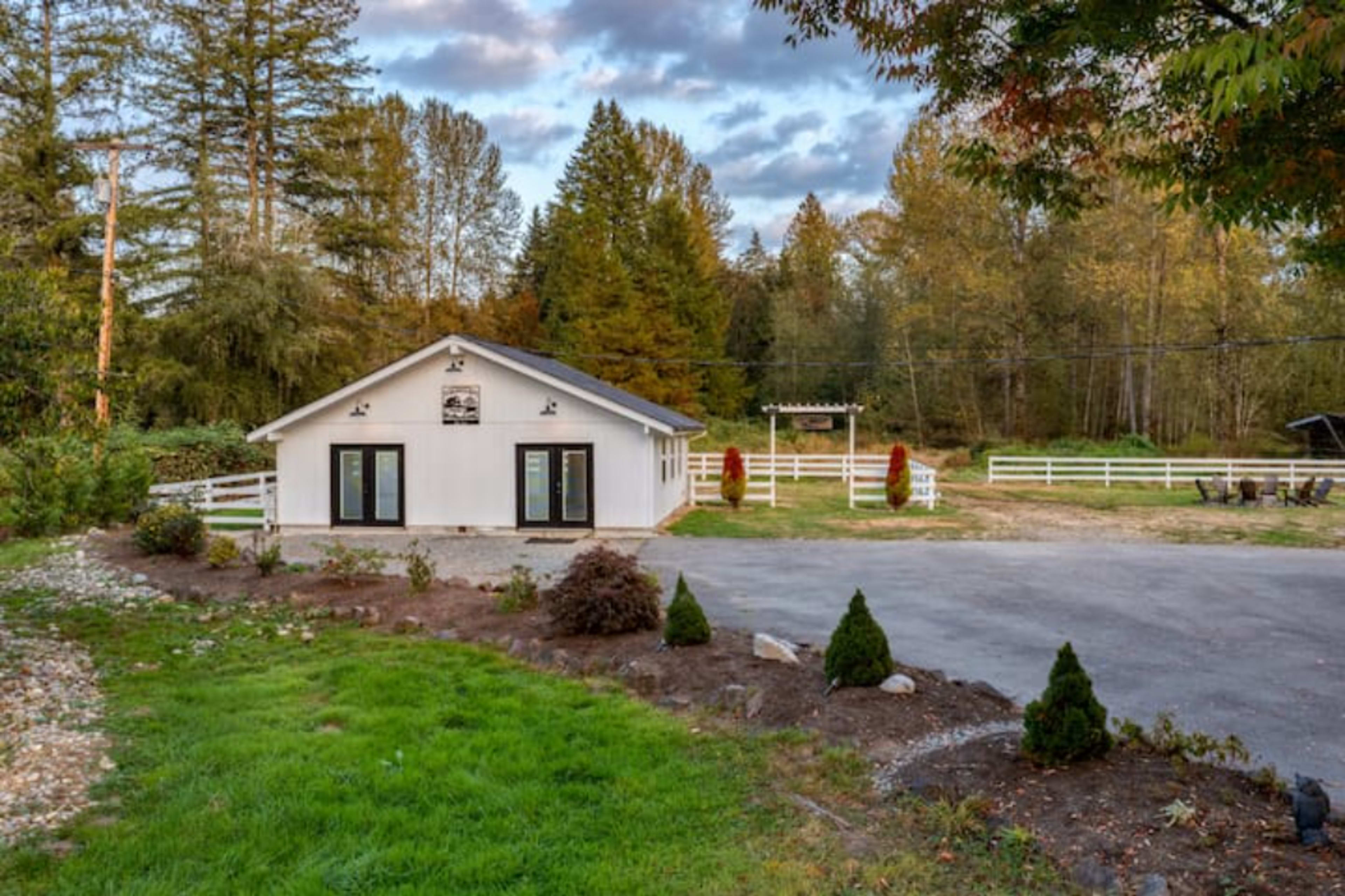 A modern white building with large windows is situated in a landscaped area surrounded by trees and a gravel driveway.