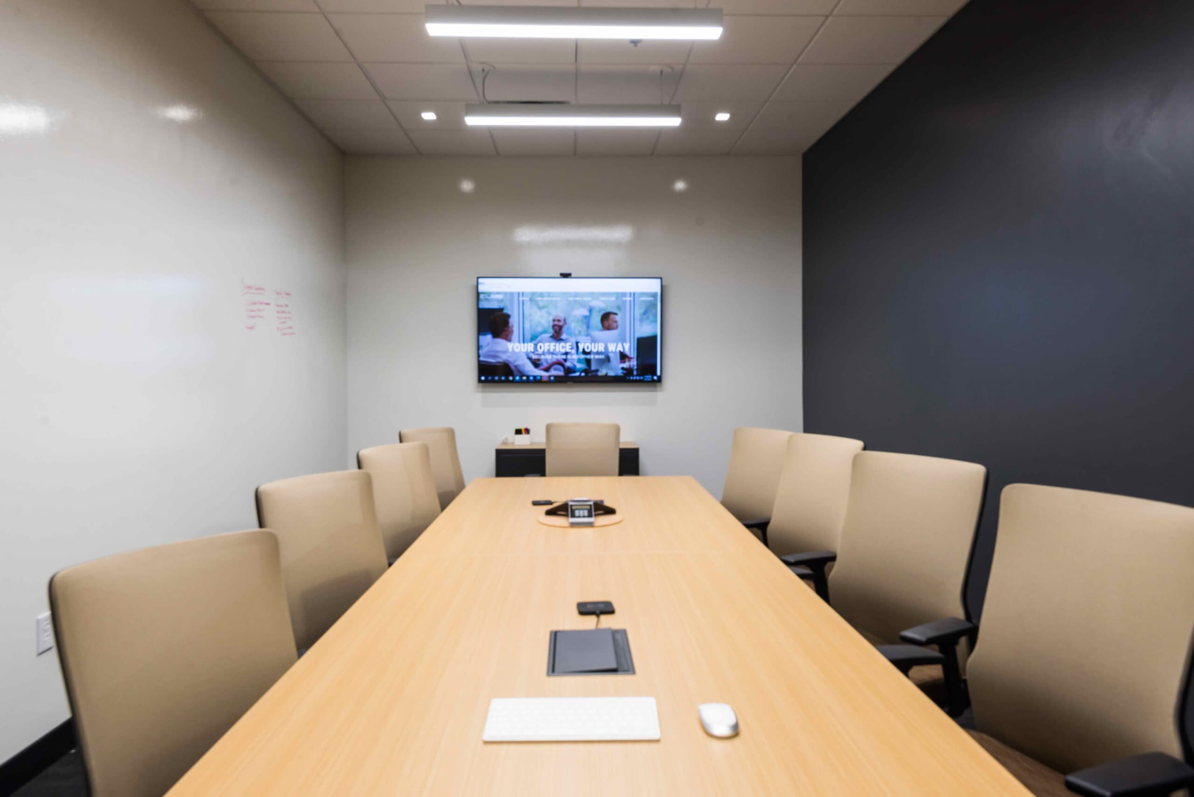 A modern conference room features a long wooden table surrounded by beige chairs, with a large screen mounted on the wall displaying a video call interface.