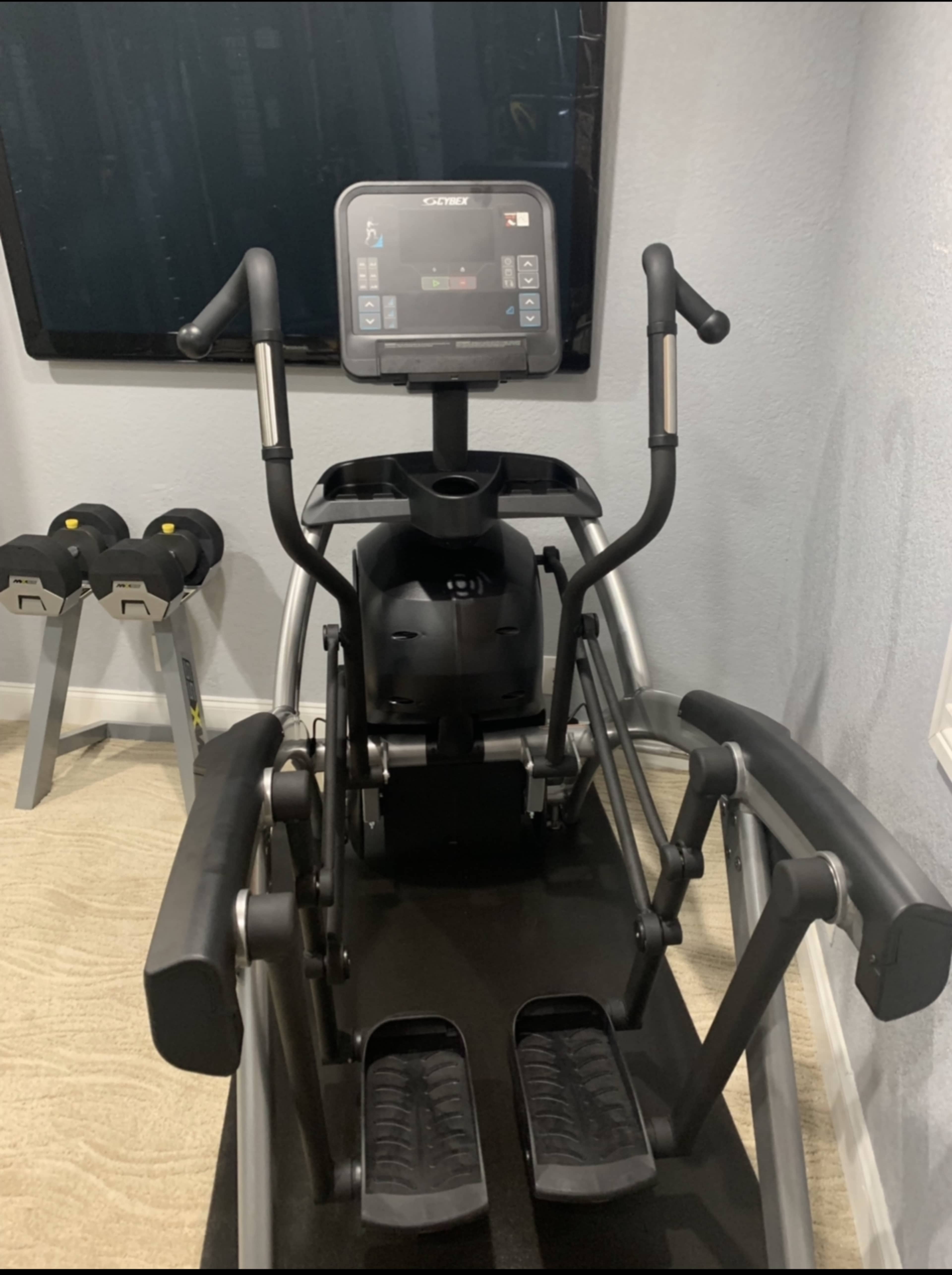 A fitness elliptical machine positioned near a wall-mounted television and a pair of dumbbells on a rack.