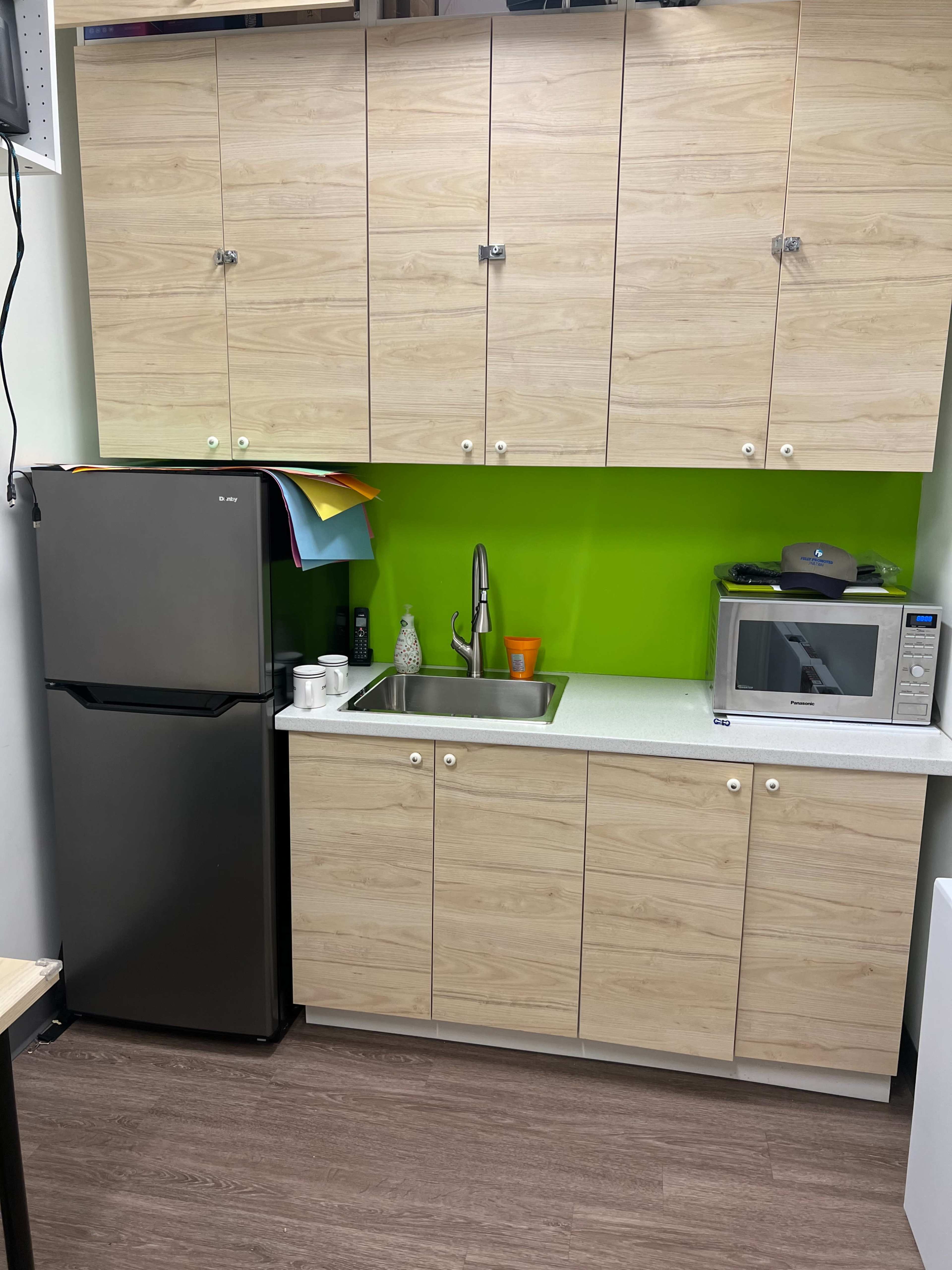 A kitchenette with a black refrigerator, a sink, a microwave, and wooden cabinets above a green wall.