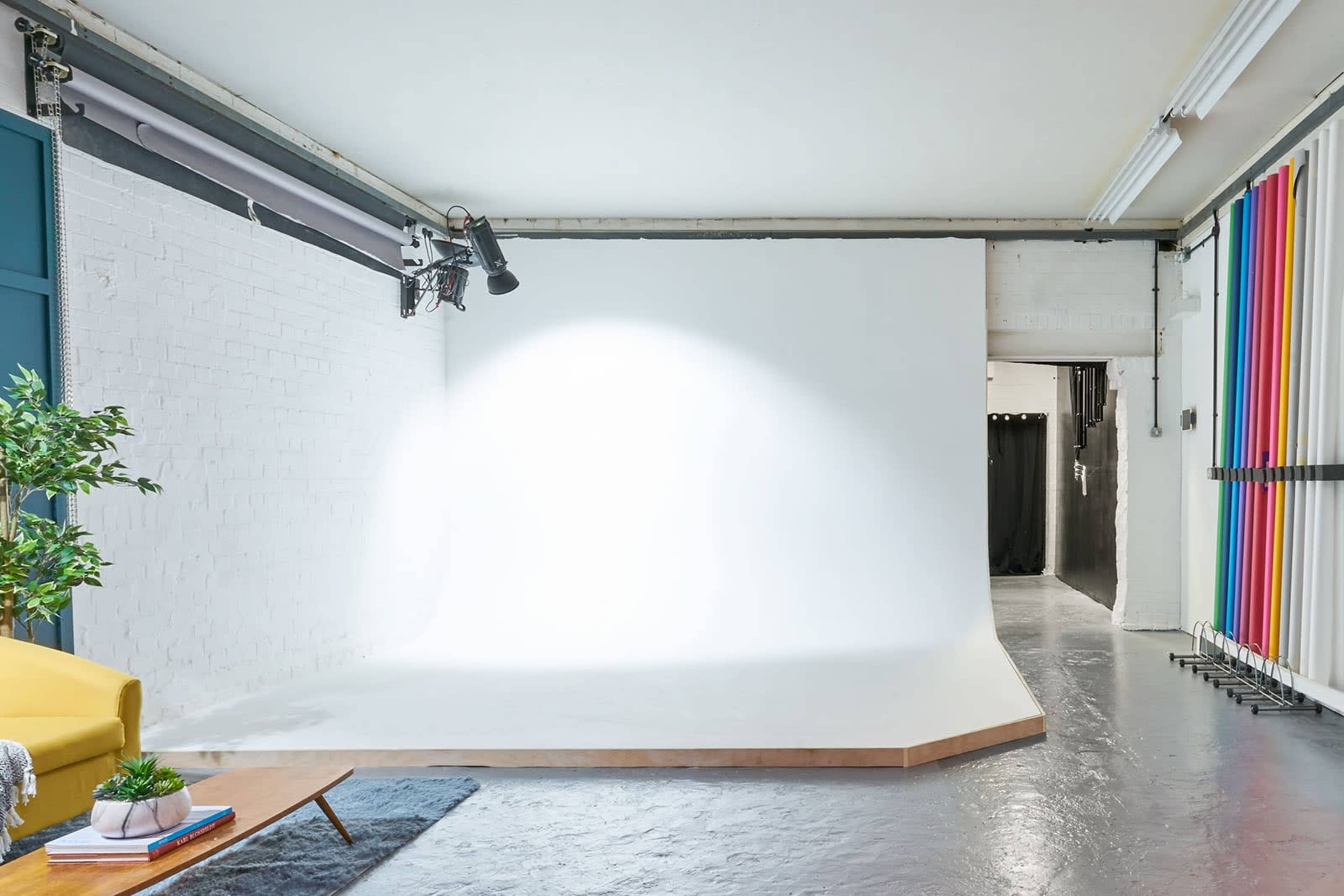 A spacious photography studio featuring a white backdrop, soft lighting fixtures, and a colorful backdrop section alongside a seating area.