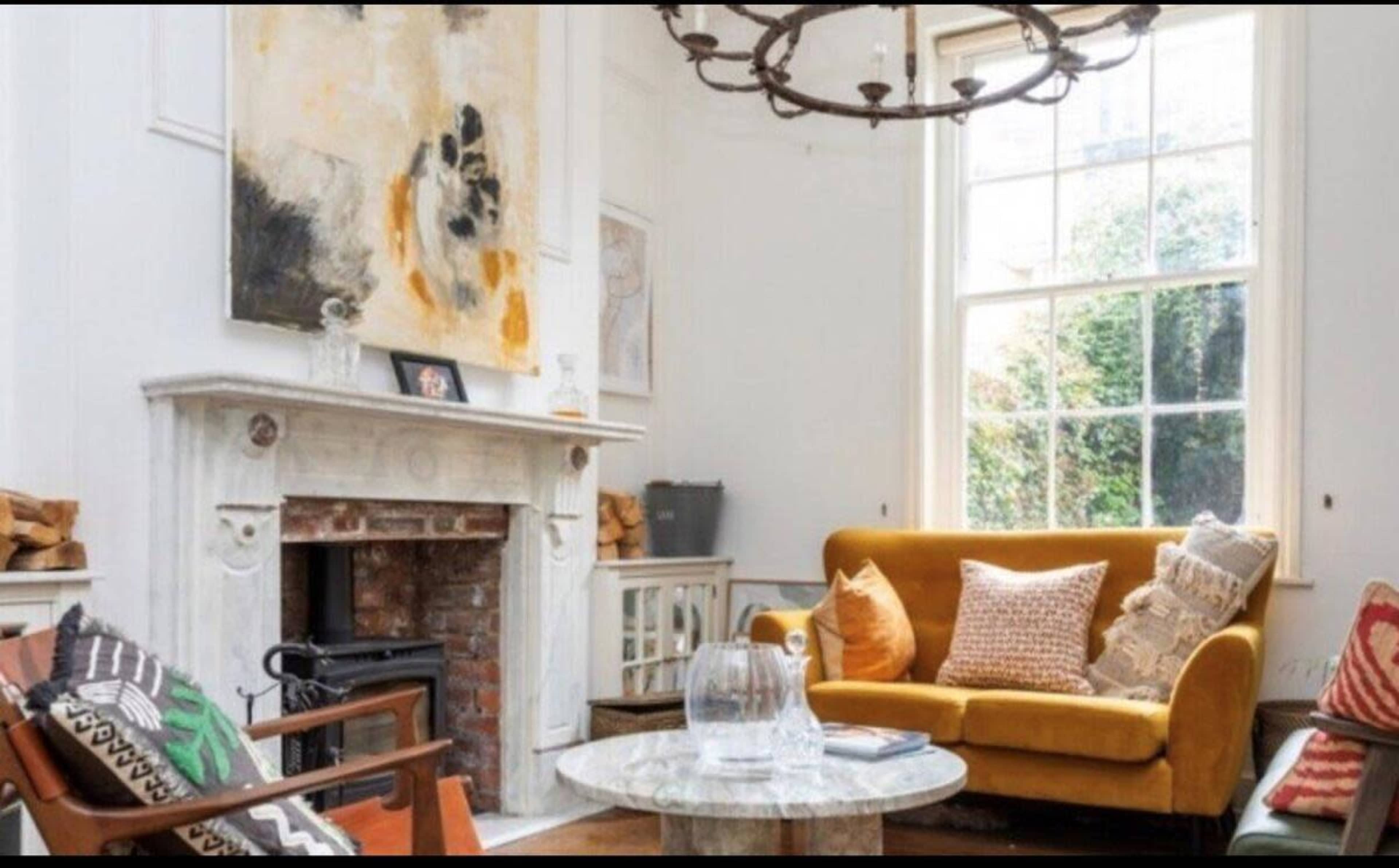A living room features a yellow sofa, a marble coffee table, and a fireplace beneath an abstract painting, with natural light streaming through a large window.