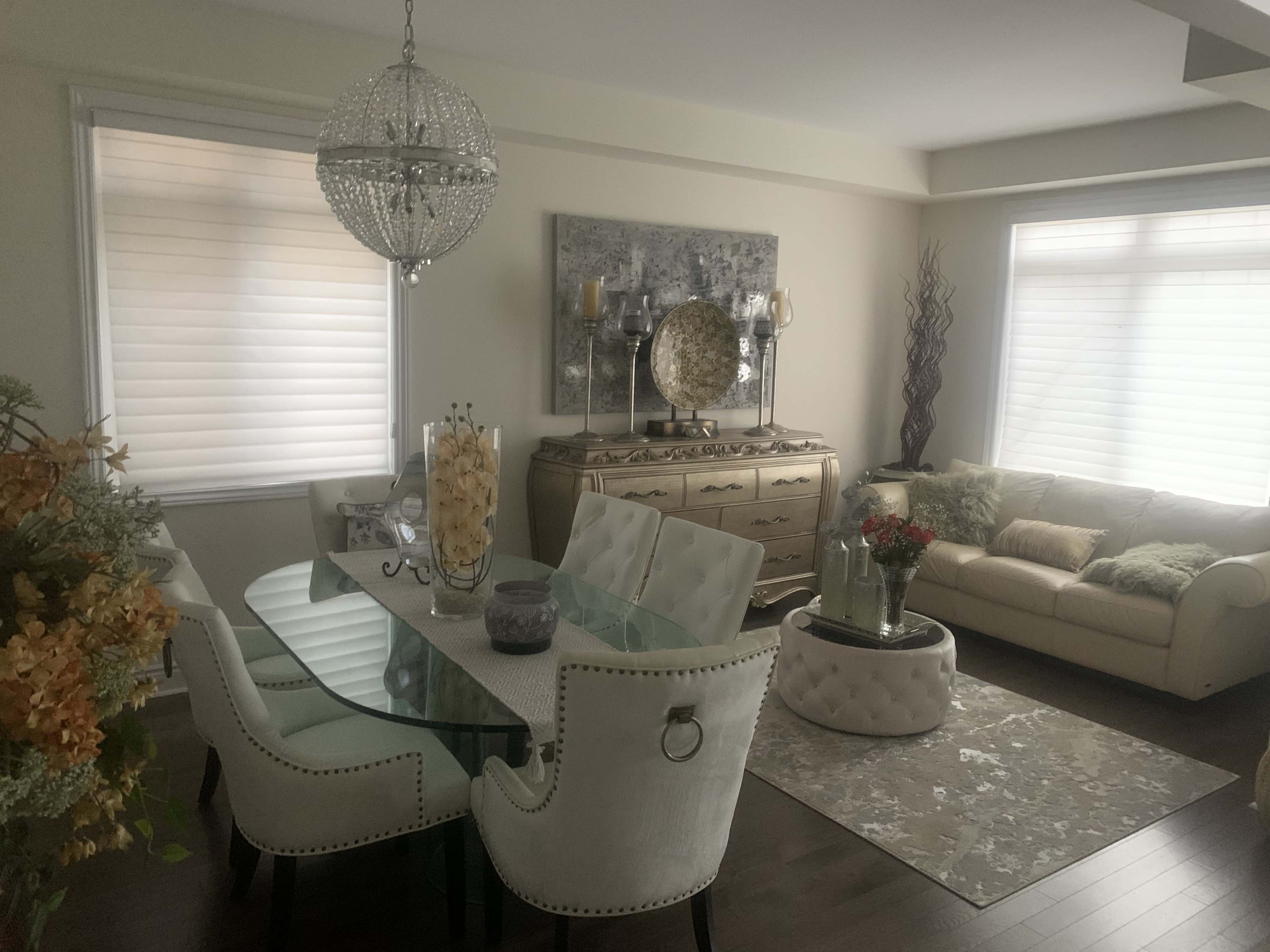 The image shows a modern living and dining area featuring a glass dining table with upholstered chairs, a cream-colored sofa, and decorative elements like a chandelier and a floral arrangement.