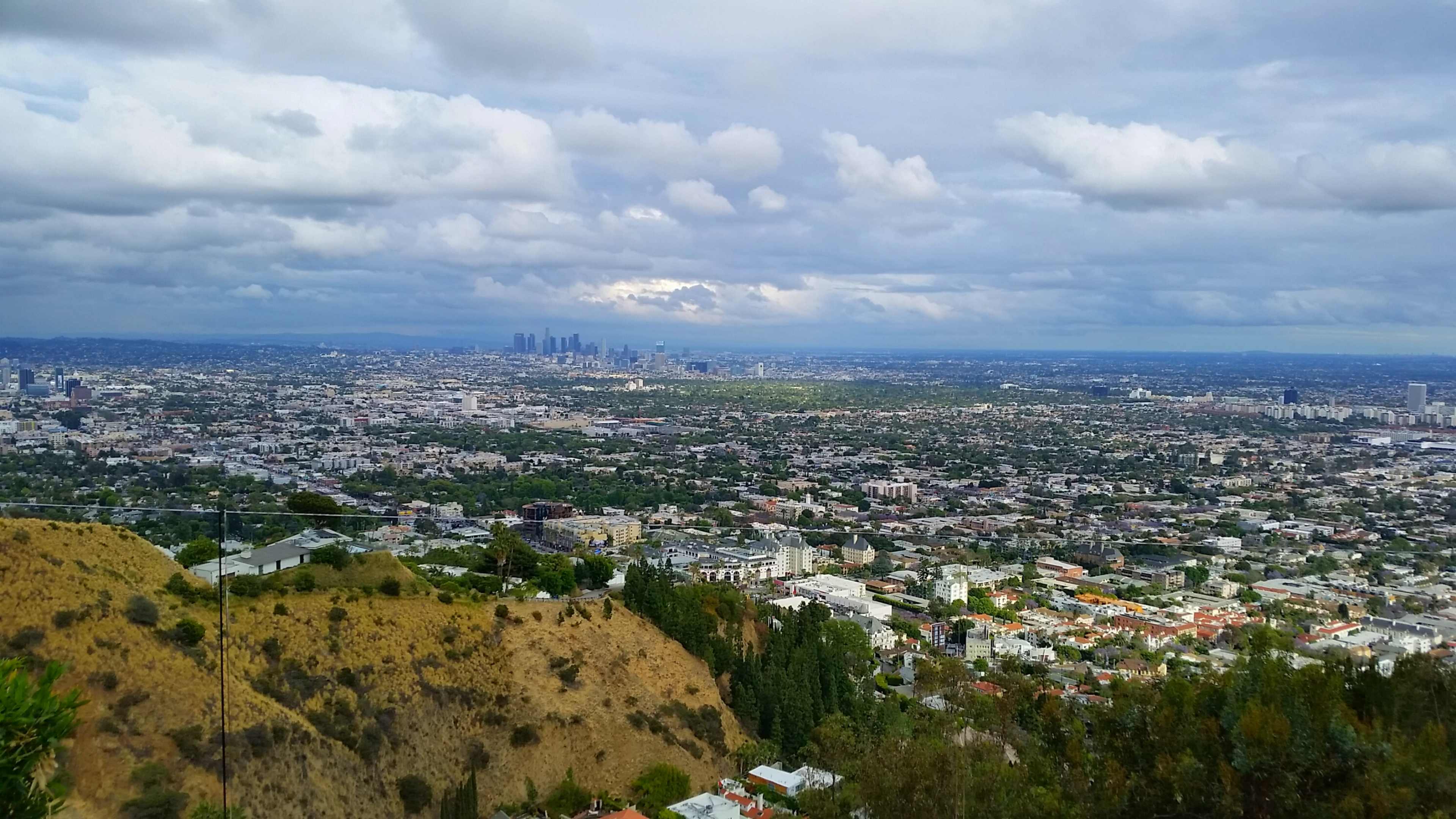 A panoramic view of a sprawling urban landscape with hills in the foreground and a city skyline in the distance under a cloudy sky.