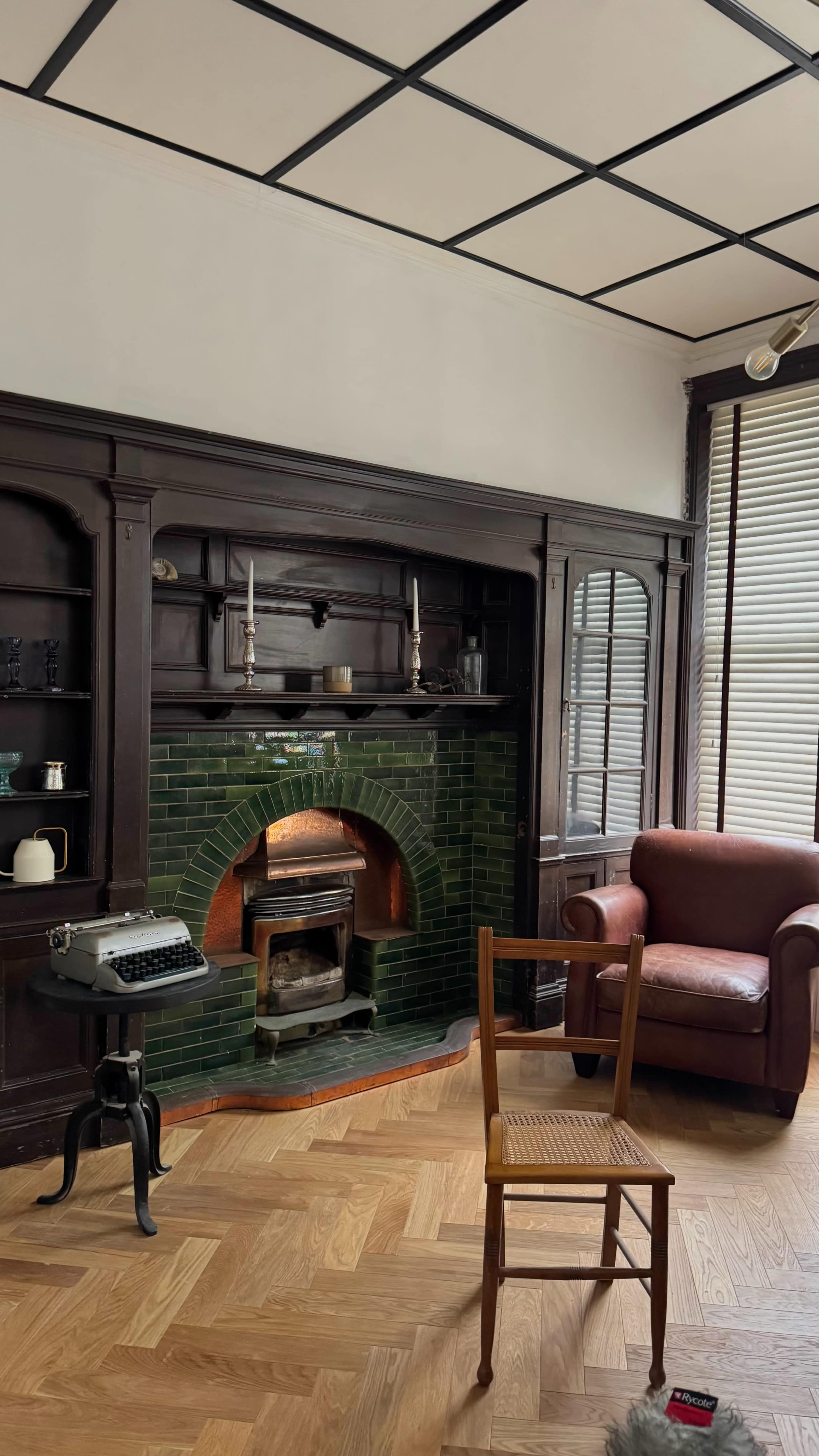 The image shows a room with a green-tiled fireplace, a vintage typewriter on a stand, a wooden chair, and a brown armchair against a wall with shelves.