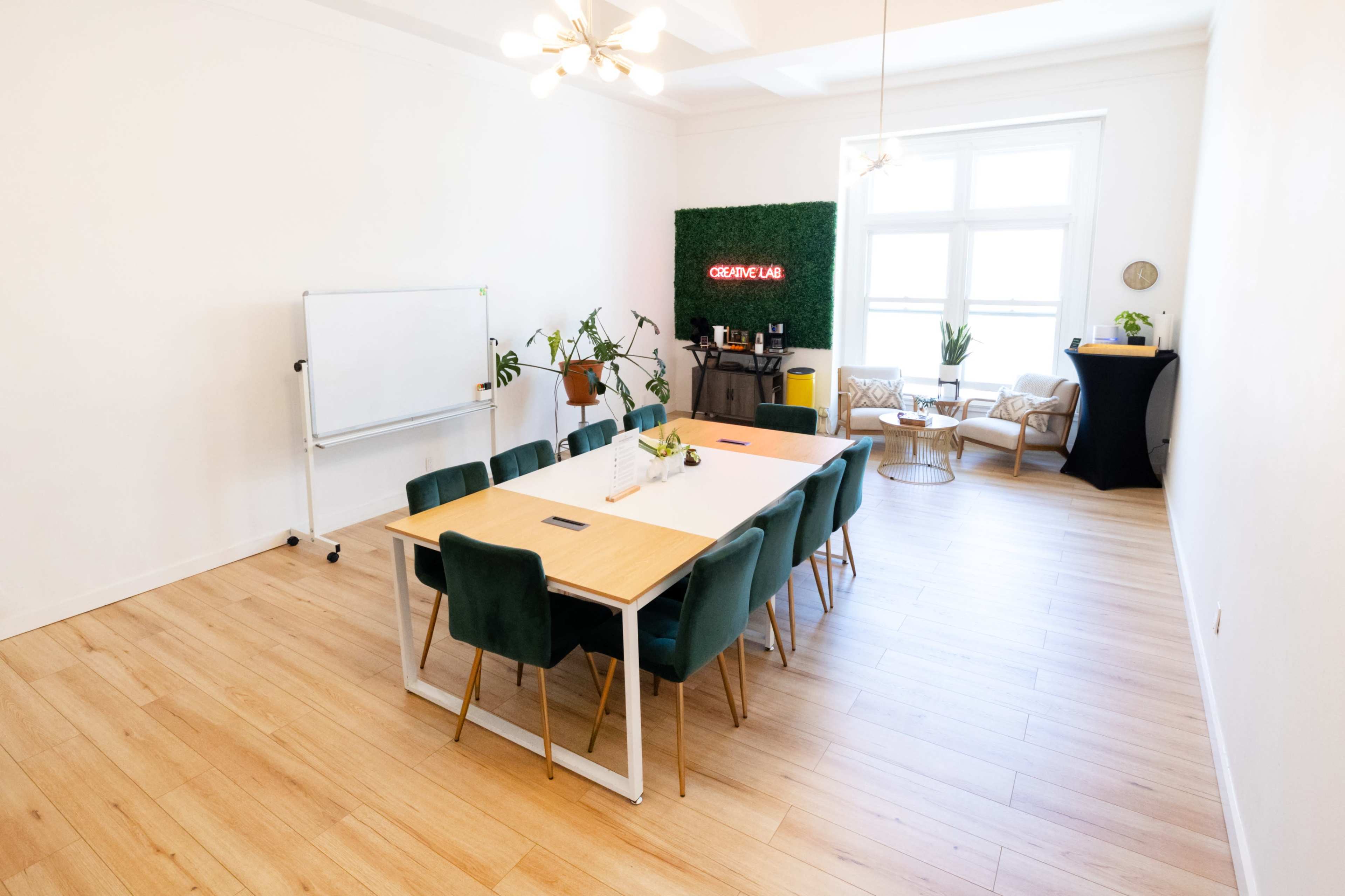 A bright meeting room features a long wooden table surrounded by green chairs, with a whiteboard on one side and a cozy seating area in the background.