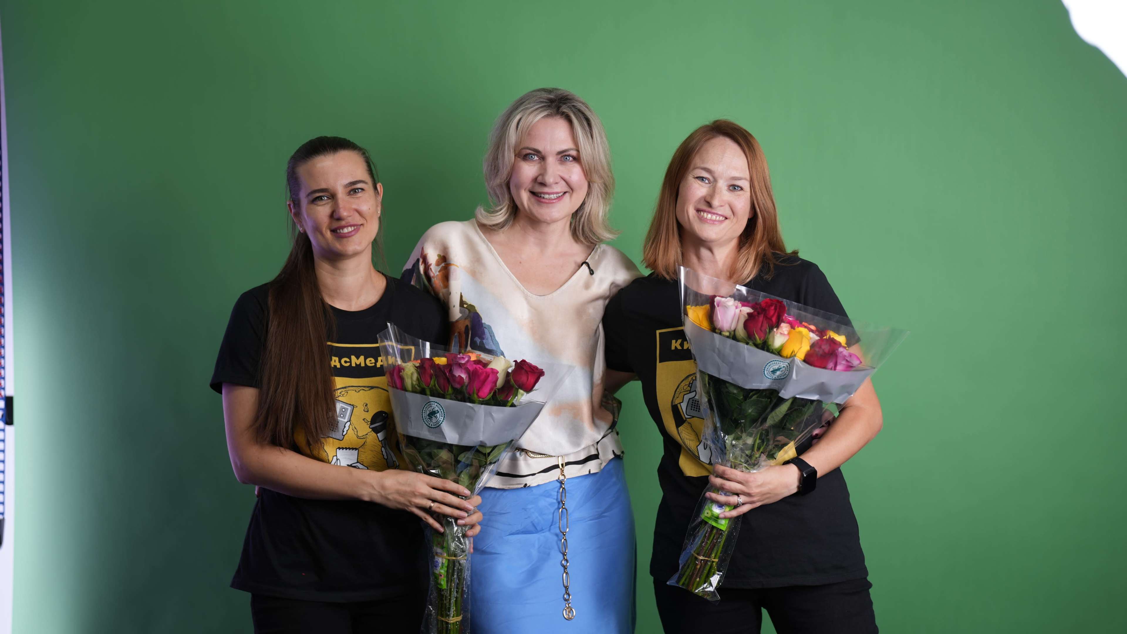 Three women, two holding bouquets of flowers and wearing matching black t-shirts, pose together in front of a green screen backdrop.