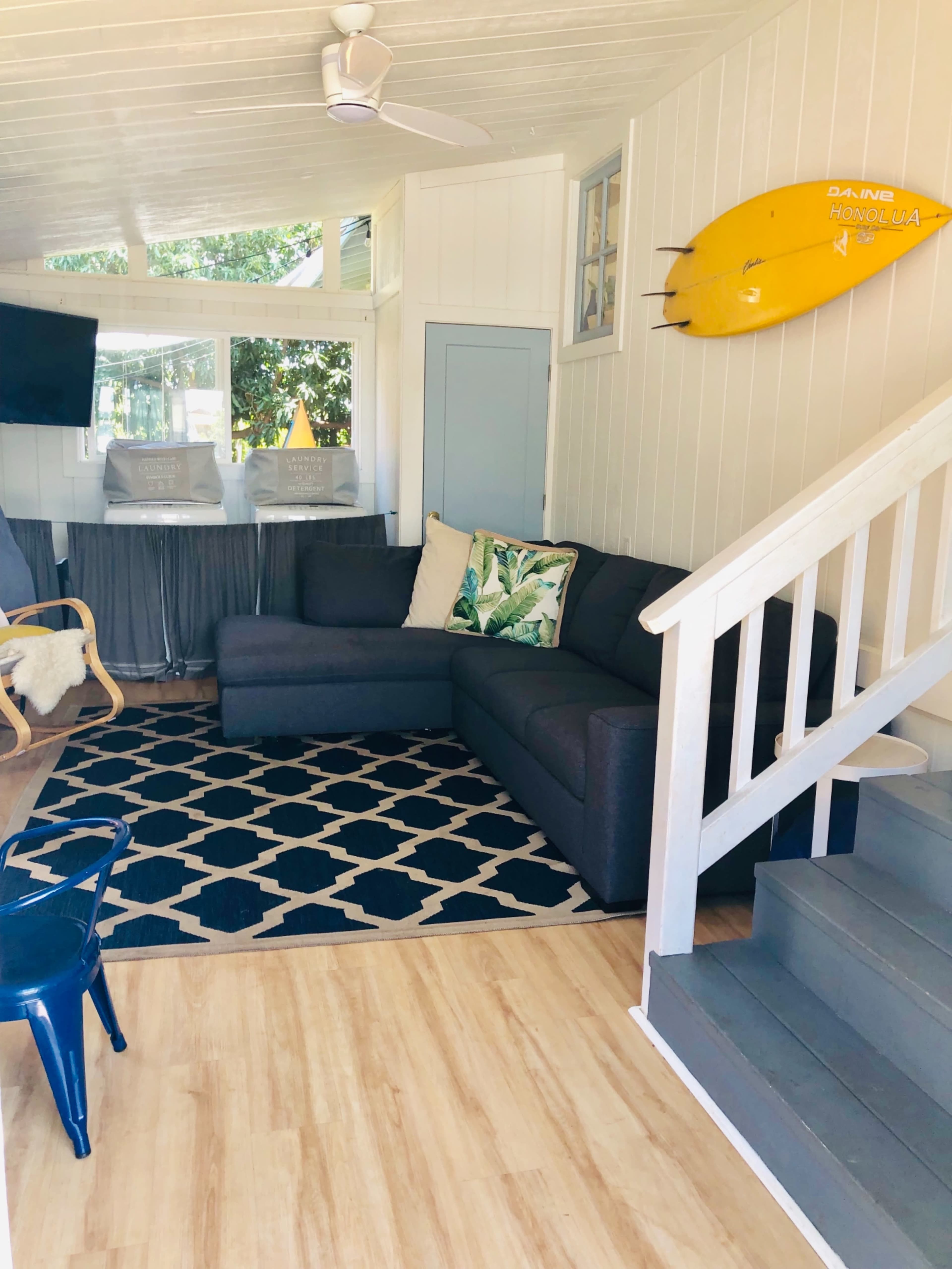 A cozy living room with a dark sectional sofa, a patterned rug, a decorative pillow, a yellow surfboard on the wall, and a staircase leading to another area.