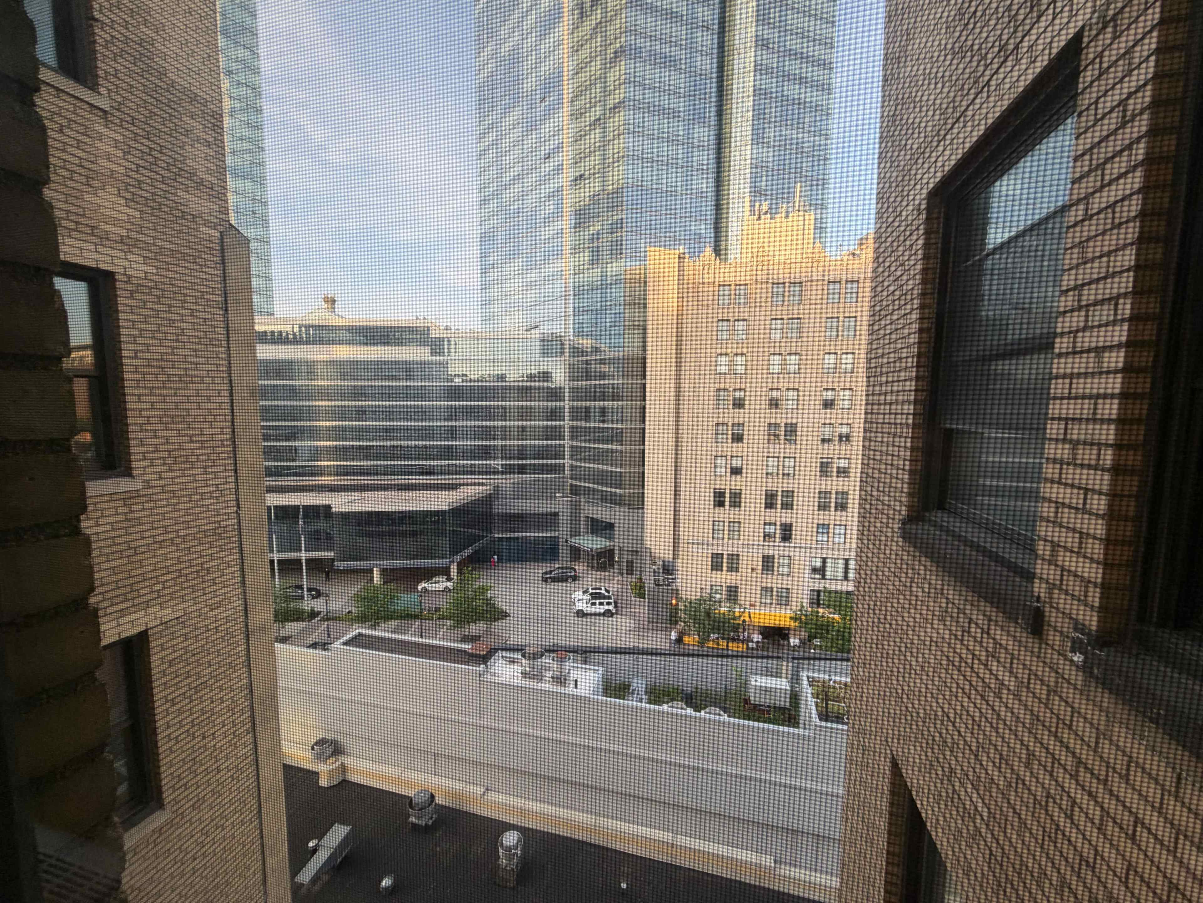 The image shows a view through a window screen of tall buildings, a courtyard, and parked cars in an urban setting.