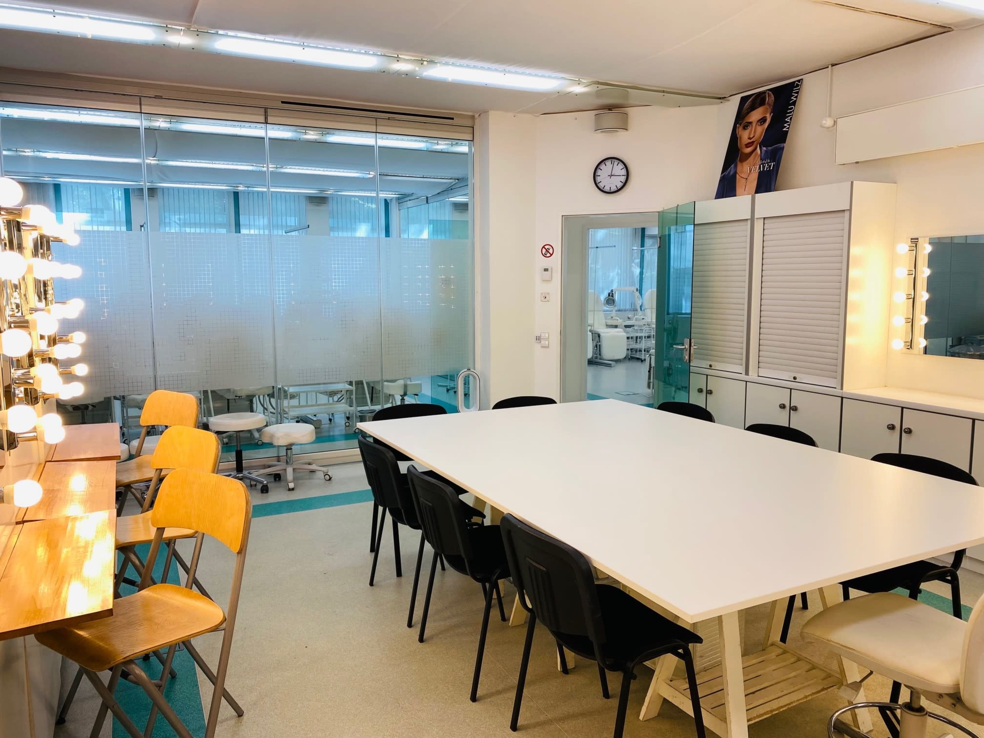 A brightly lit room with a large table and several chairs, alongside a vanity area with mirrors and lights, and a background of medical equipment visible through glass partitions.