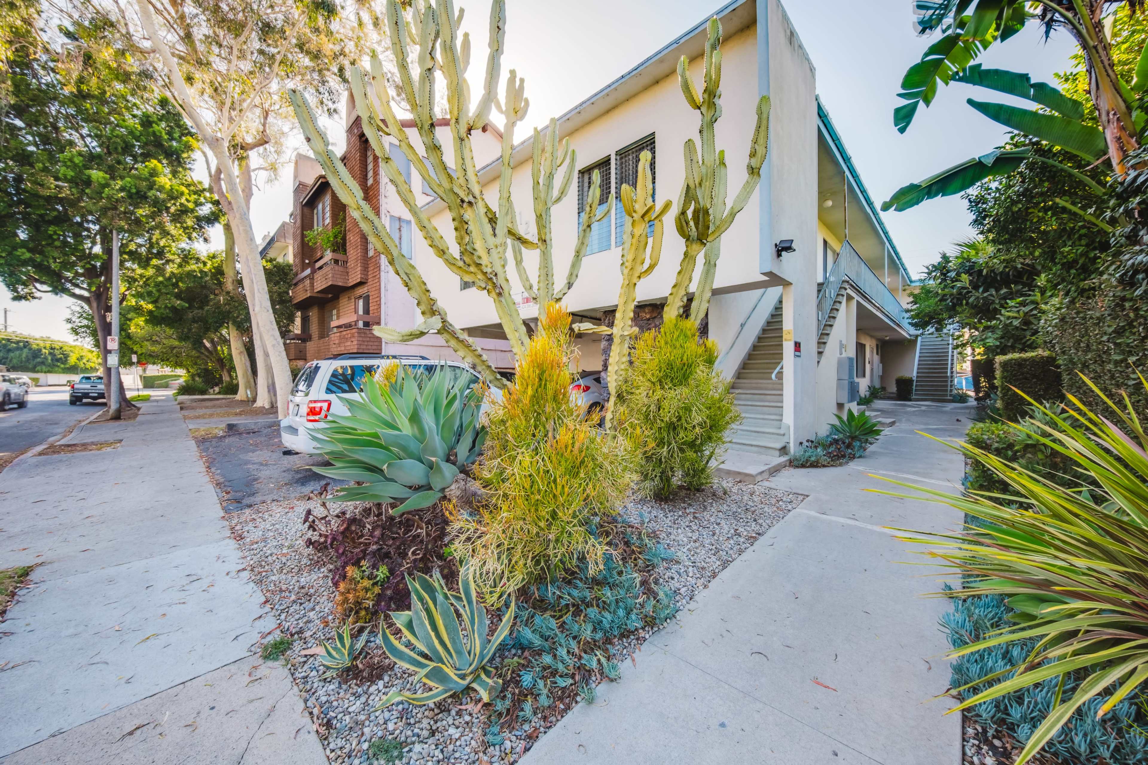The image shows a residential building with a landscaped front area featuring various plants, including cacti and succulents, alongside a sidewalk and parked cars.