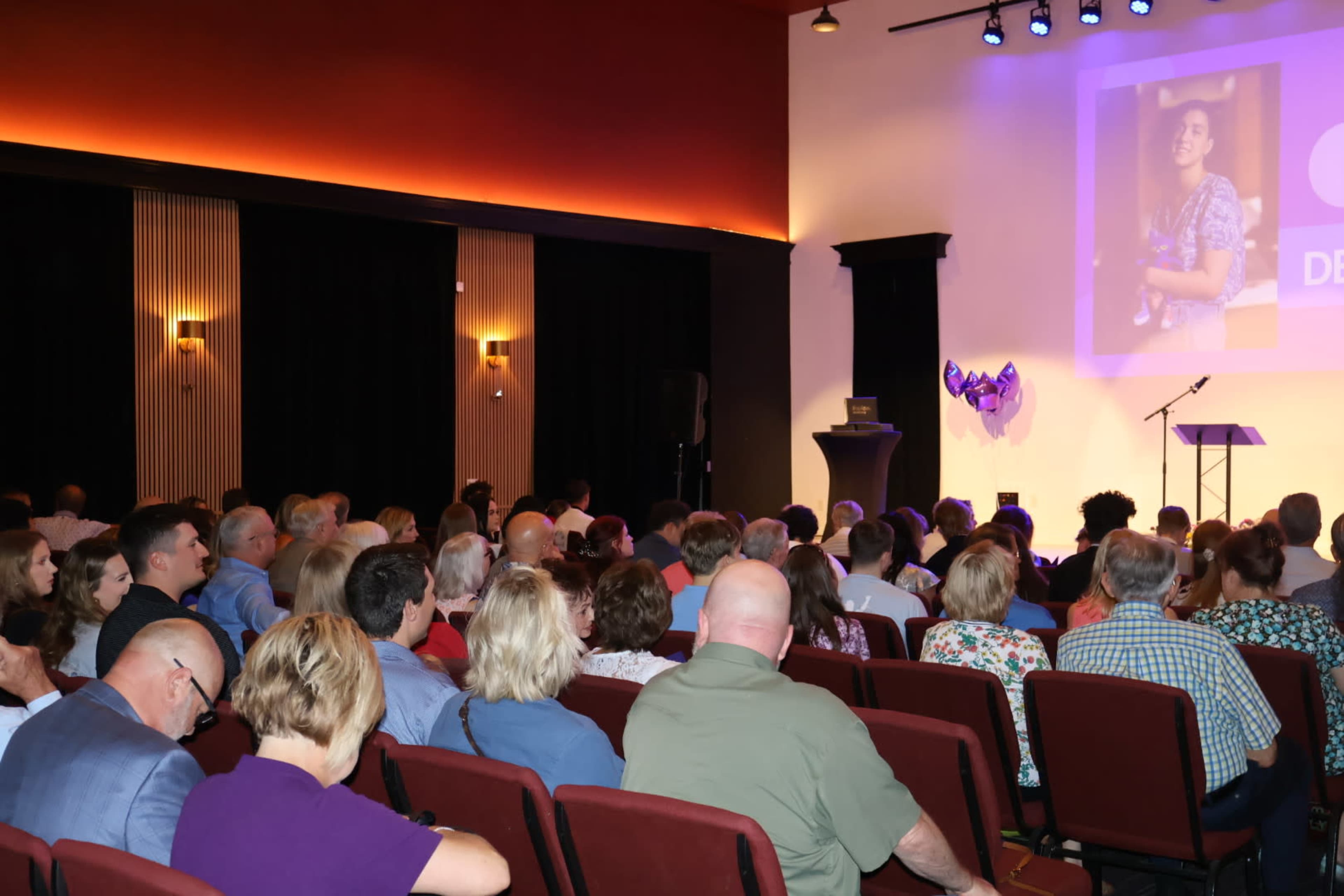 A large audience is seated in a dimly lit room, facing a stage with a presentation screen and a podium.