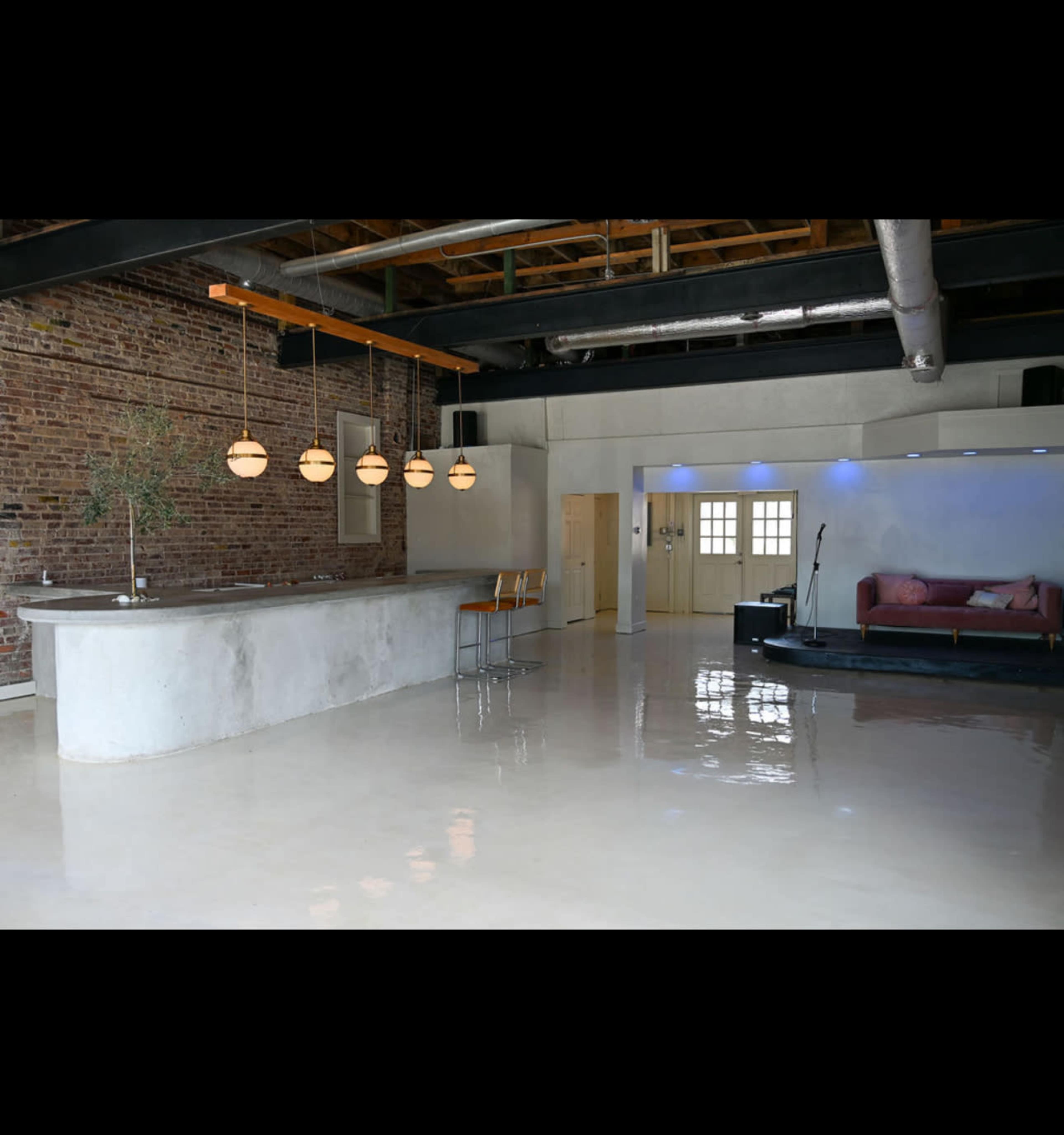 The image shows a spacious interior featuring a modern kitchen island with pendant lights, polished concrete floors, and a sitting area with a couch against a brick wall.