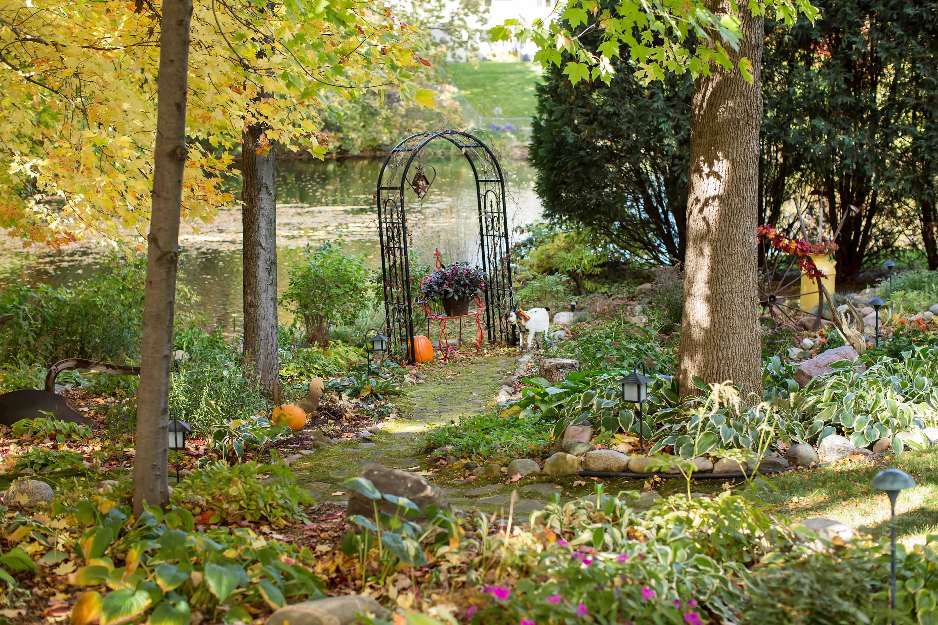 A stone path leads through a garden arch adorned with flowers, bordered by trees and a pond in the background.