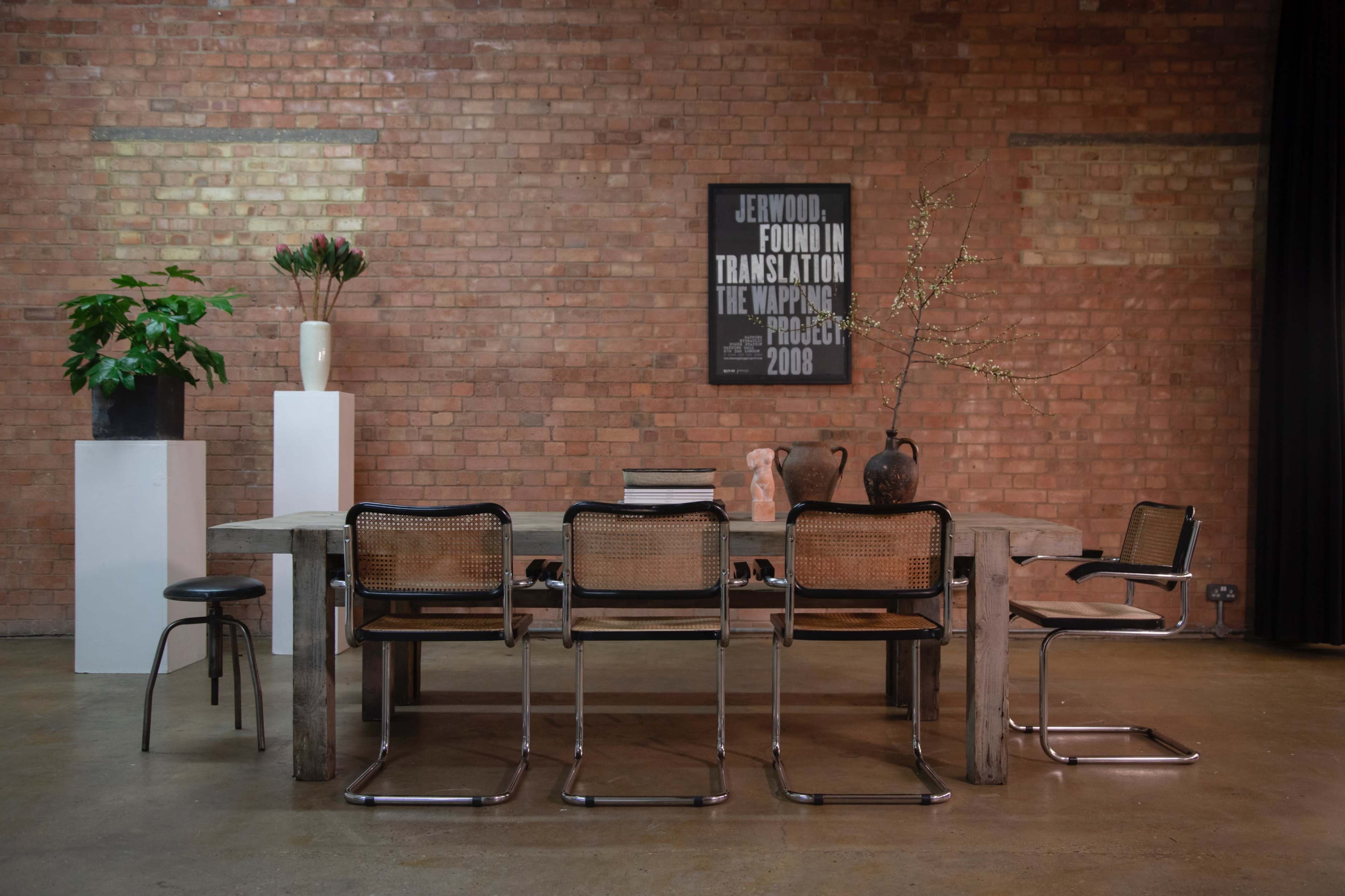 A wooden dining table is surrounded by metal chairs in an industrial-style room with exposed brick walls and various decorative plants and vases on display.