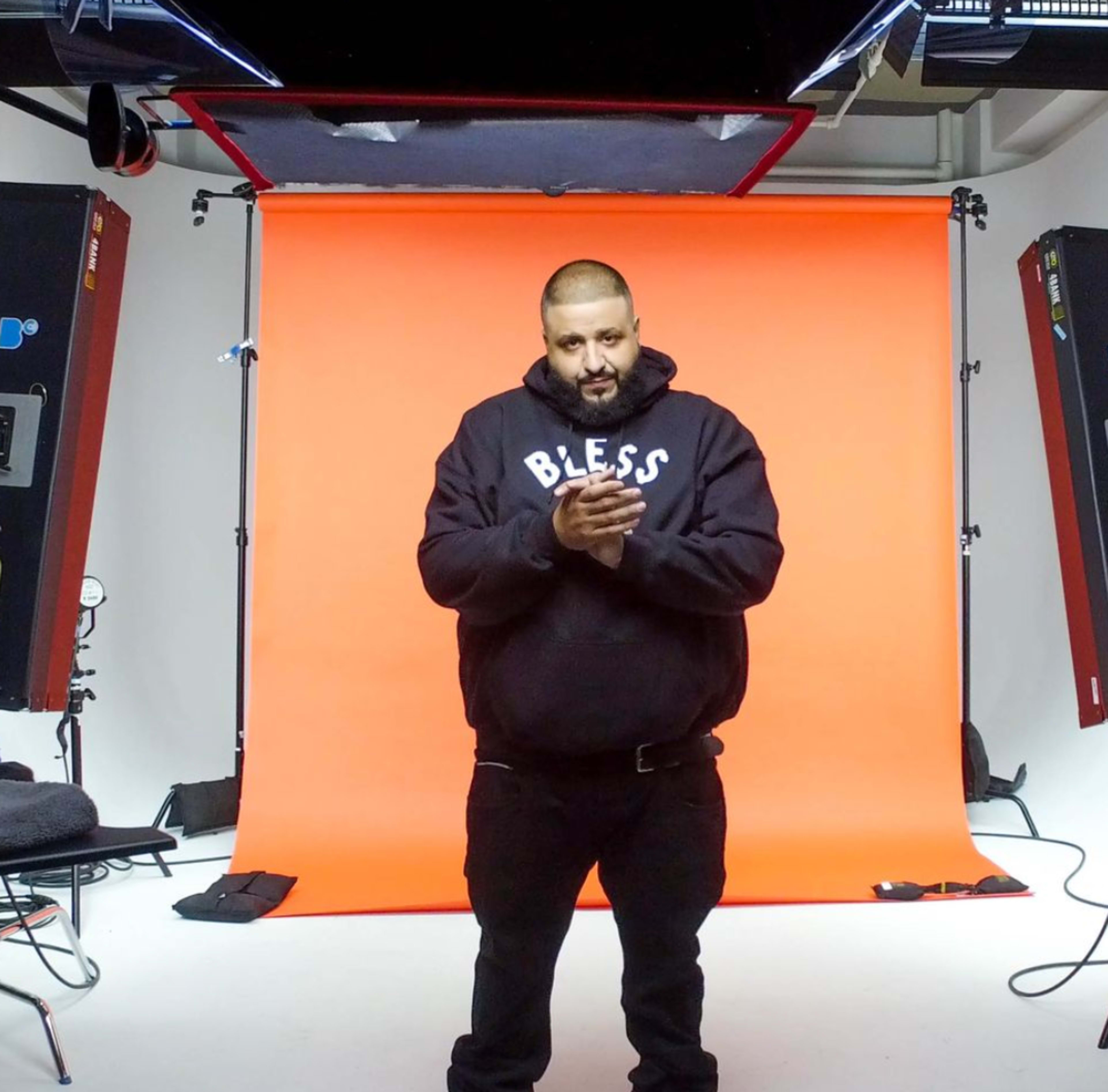 A man stands in front of an orange backdrop, clapping his hands, with studio lights positioned around him.
