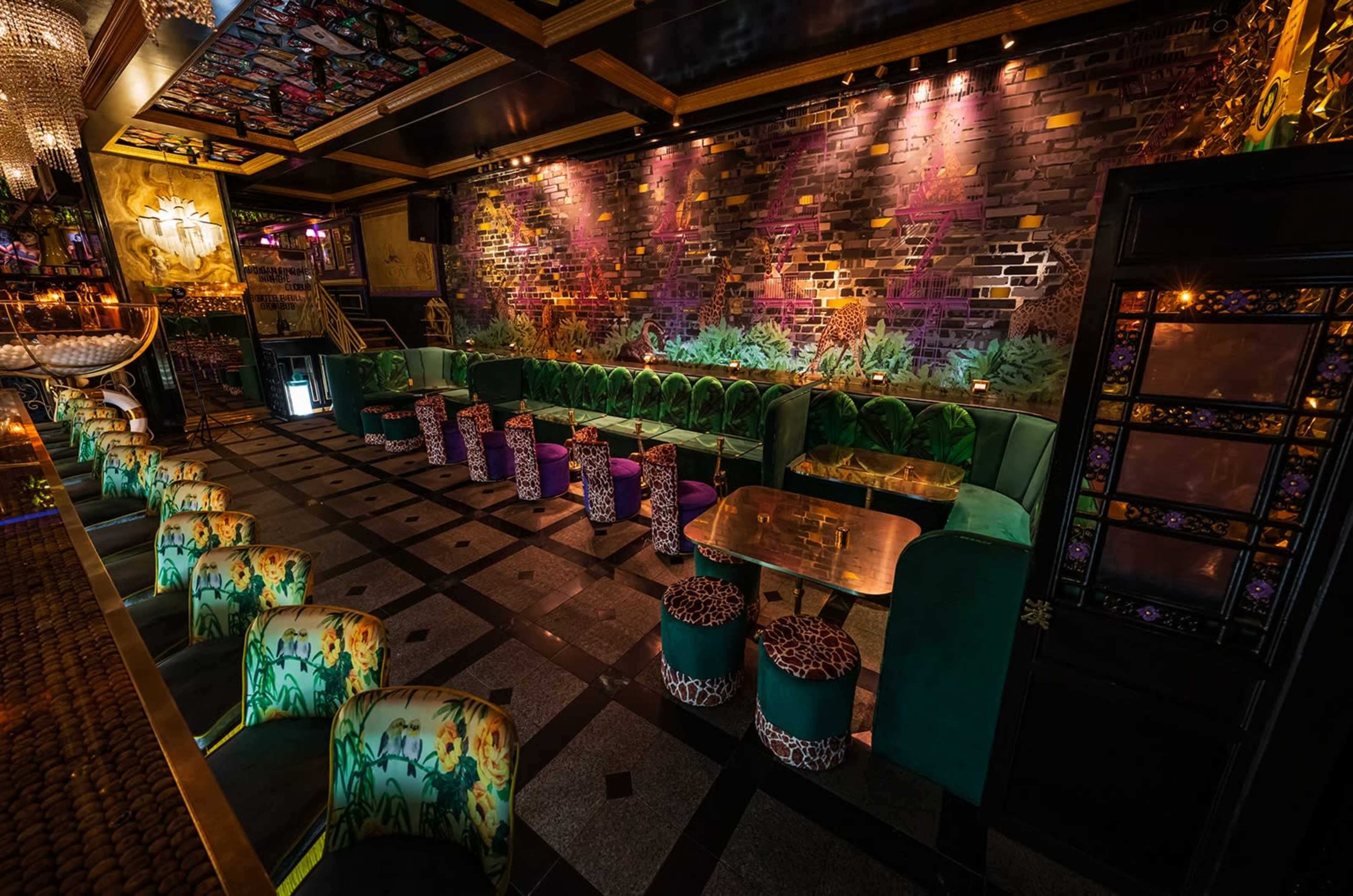 The image depicts a stylish bar area with vibrant green seating, ornate stools, and an artistic brick wall adorned with colorful murals.