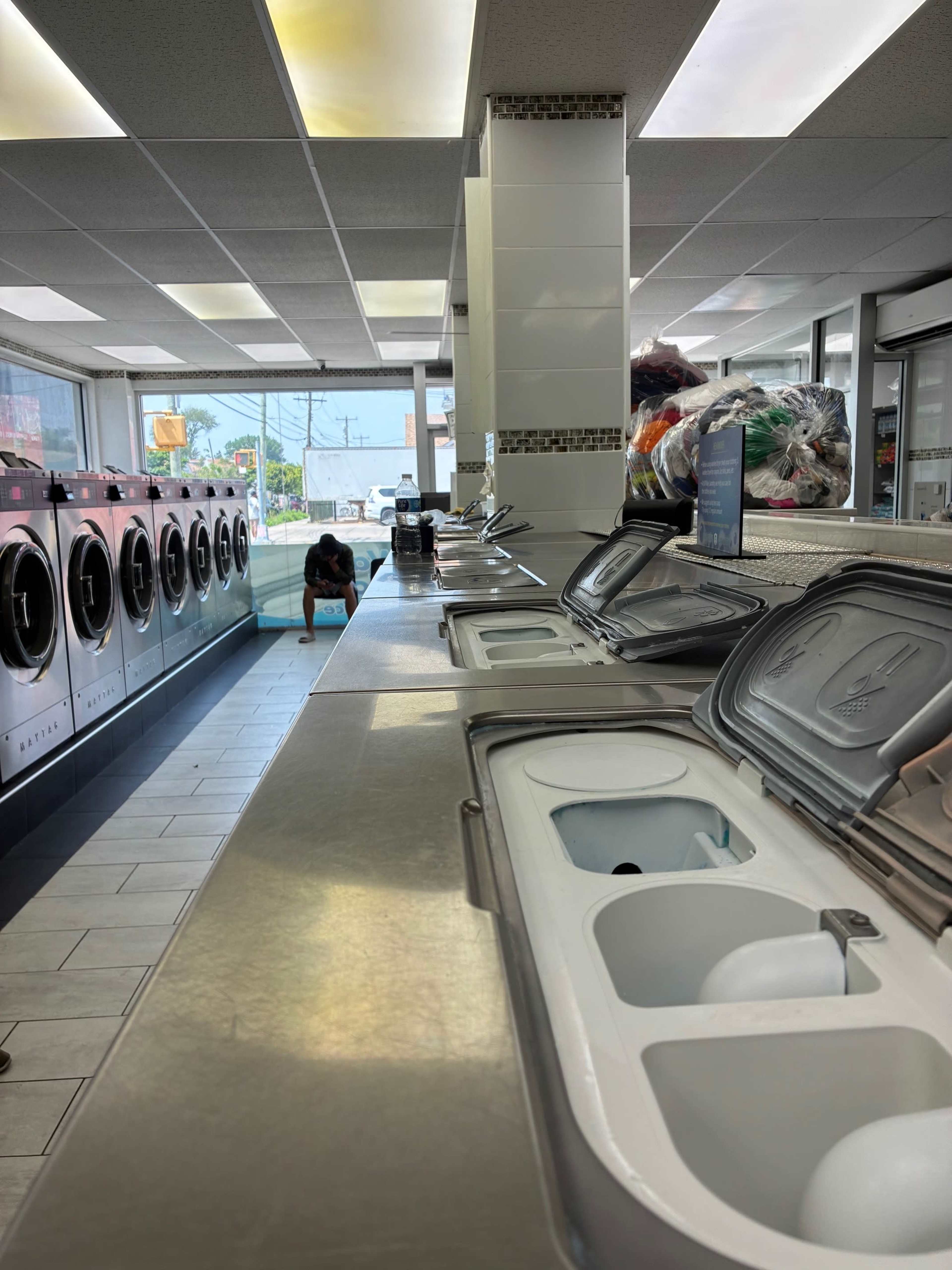 The image shows a laundromat interior with rows of washing machines and a woman crouching near the end of a counter.