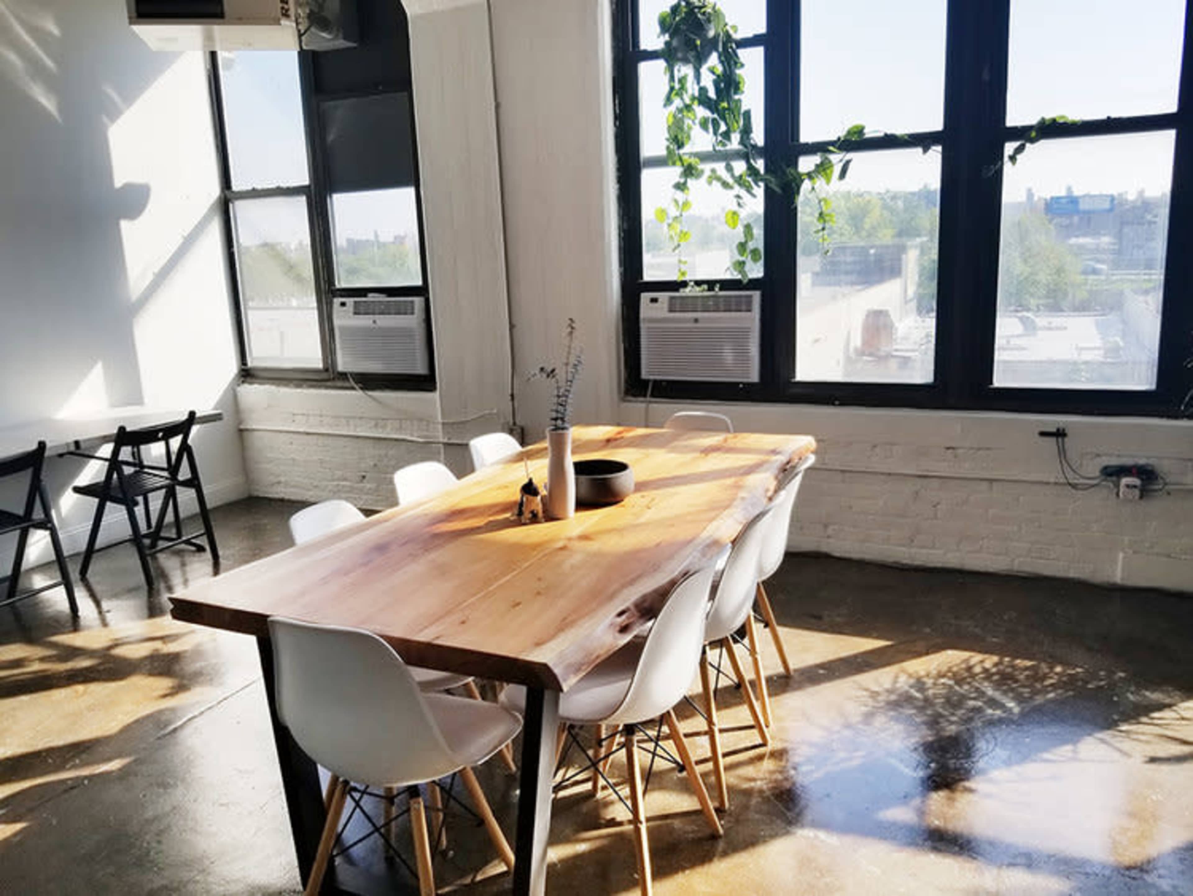A large wooden table with white chairs is set in a well-lit room featuring tall windows and indoor plants.