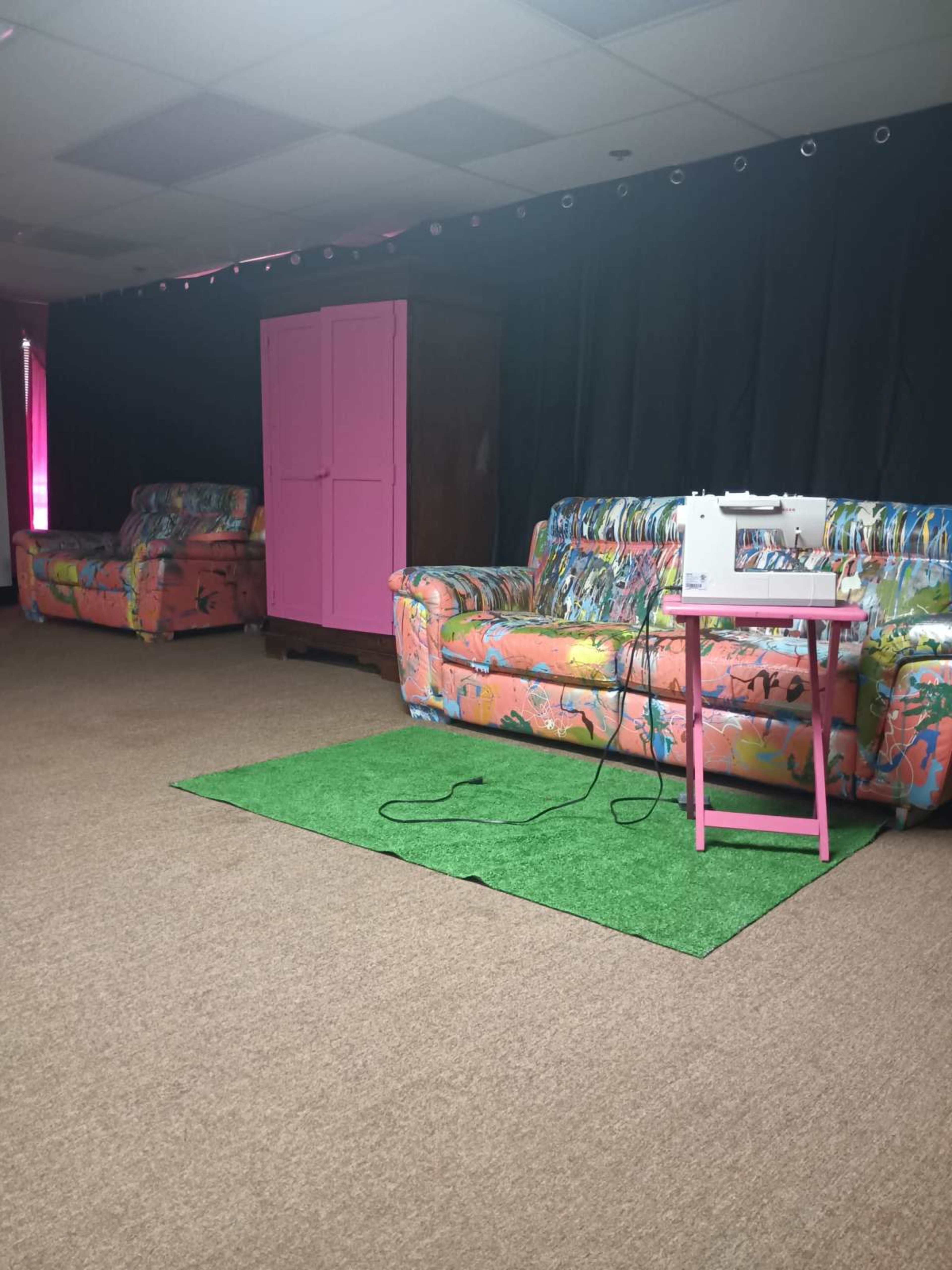 The image shows a room with two colorful couches, a pink side table with a sewing machine, and a dark backdrop.