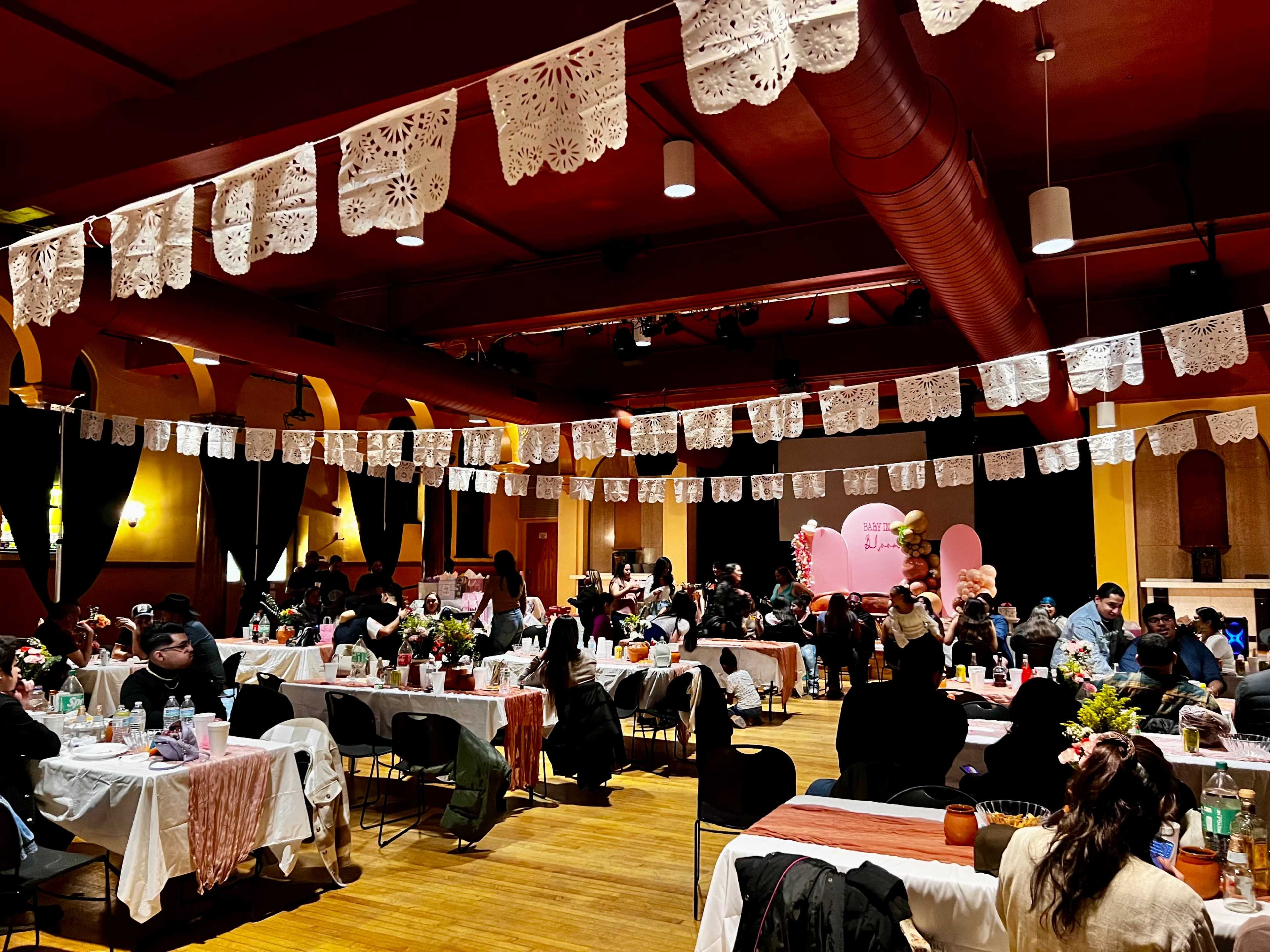 A vibrant event space is filled with tables decorated with flowers and tableware, while colorful papel picado banners hang above attendees engaging in conversation.