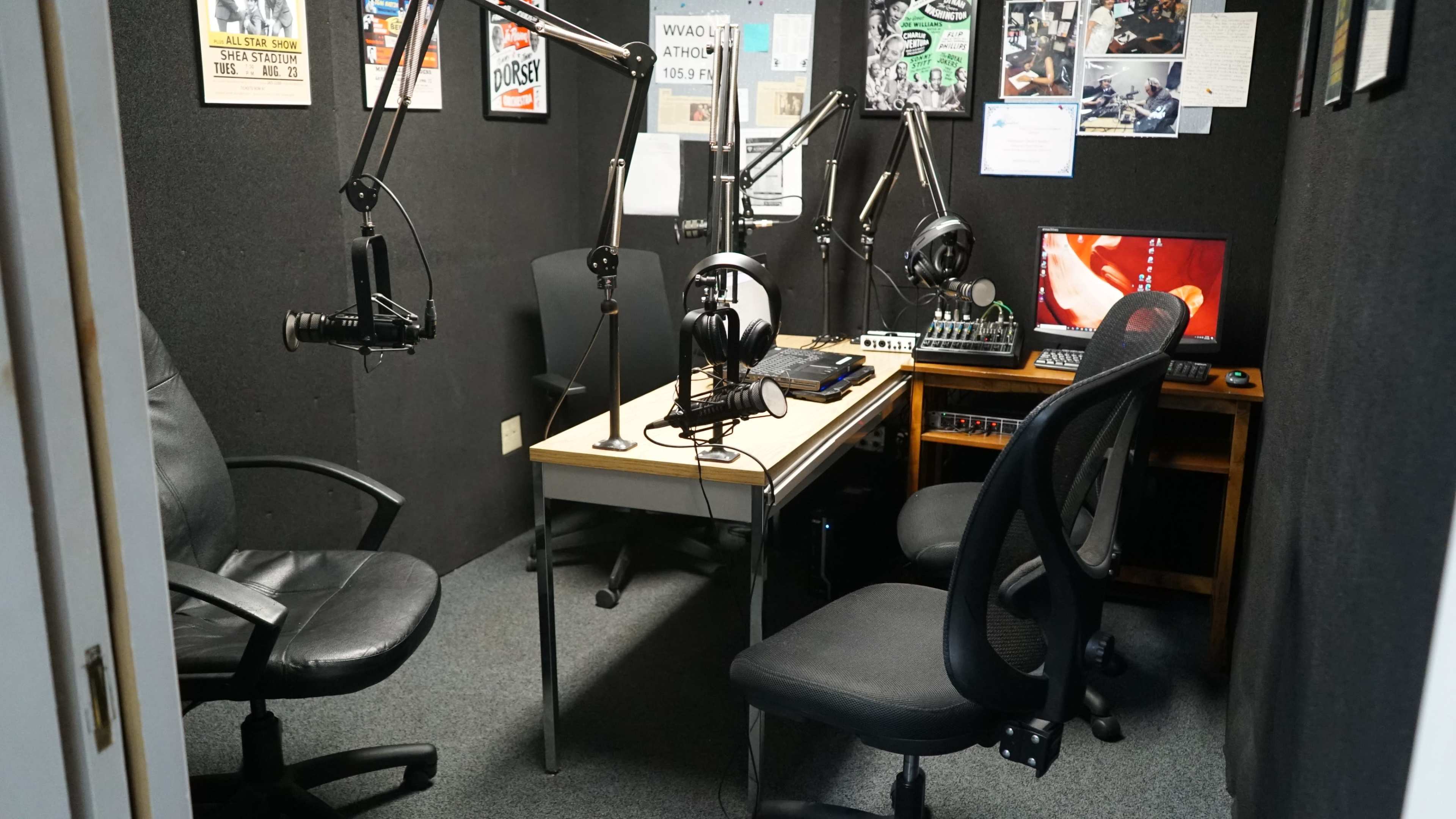 The image shows a small recording studio with two chairs, a table equipped with microphones, headphones, and audio equipment on display.
