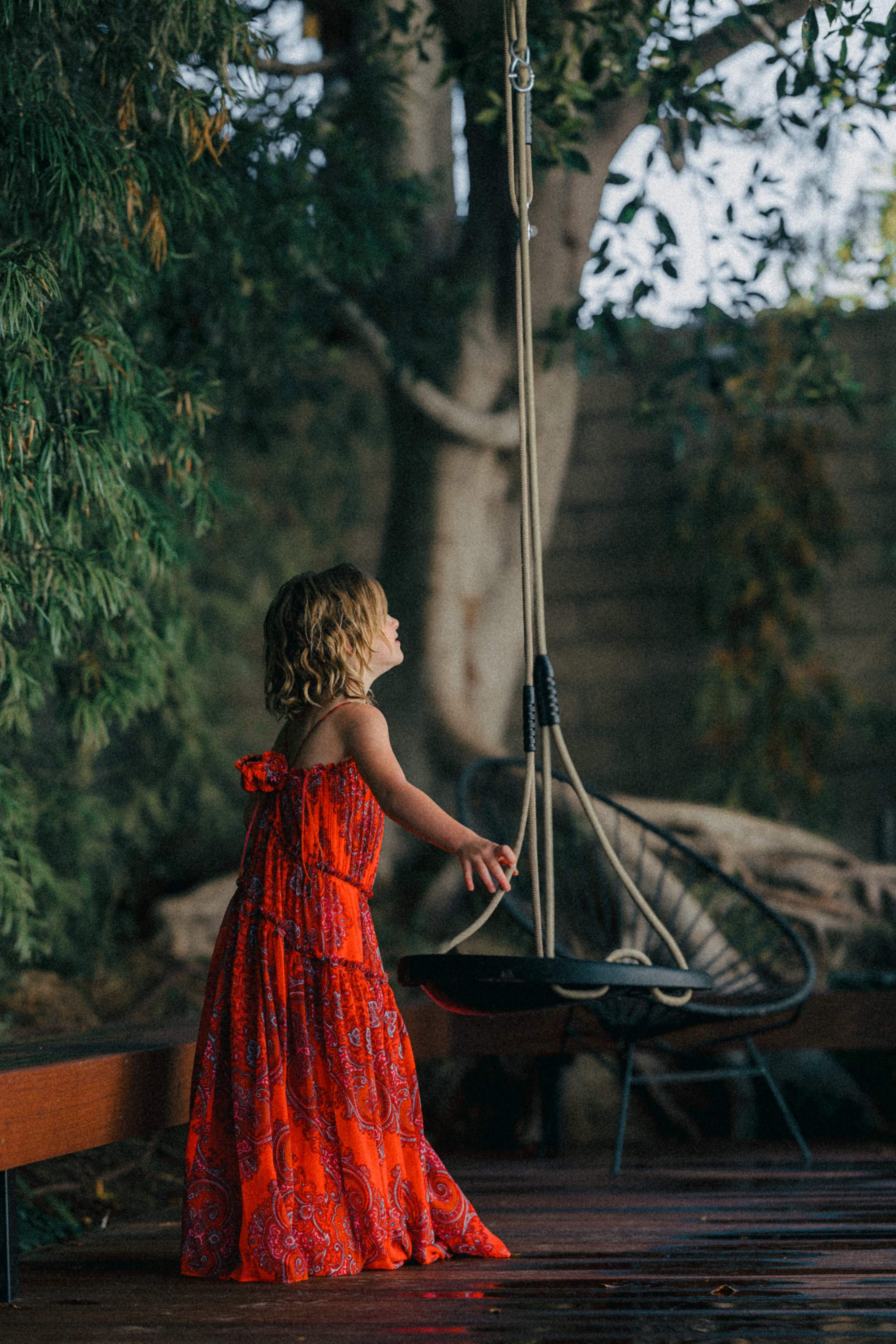 A young girl in a vibrant red dress stands on a wooden deck, reaching for a swing that hangs from a tree.