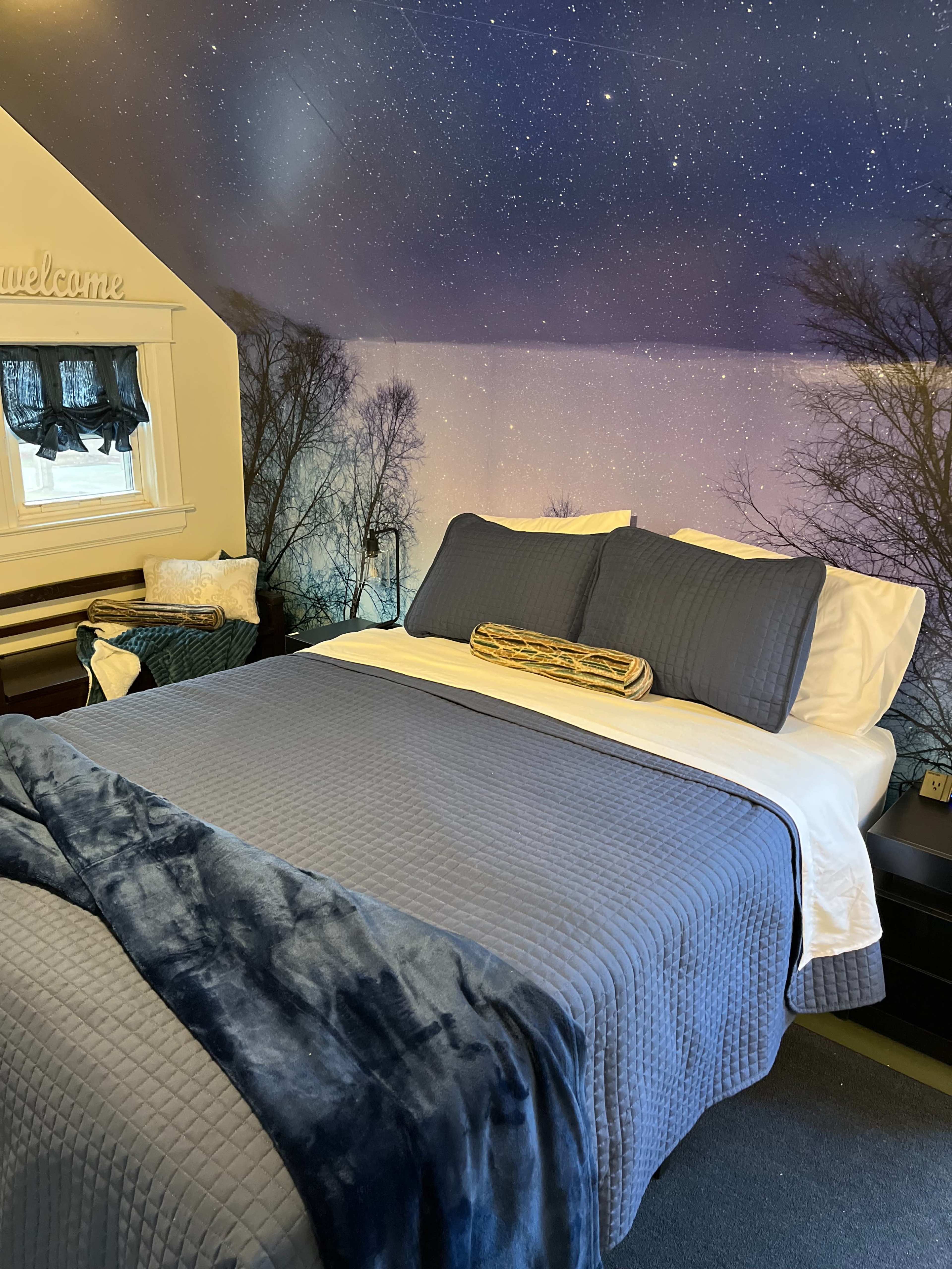 A neatly made bed with dark blue bedding in a room featuring a starry night mural on the wall.