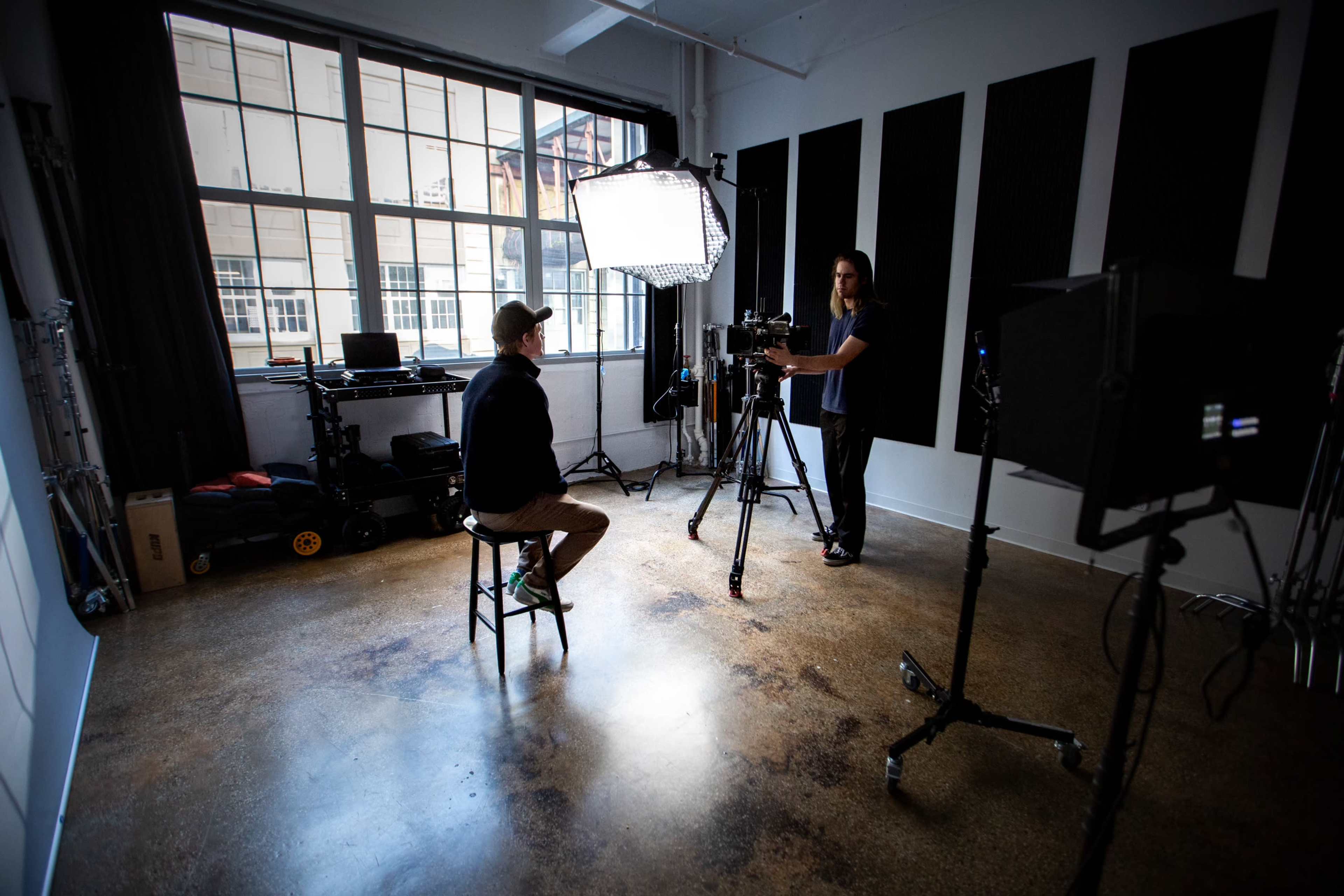 A person sits on a stool in a studio while another person adjusts a camera positioned on a tripod.