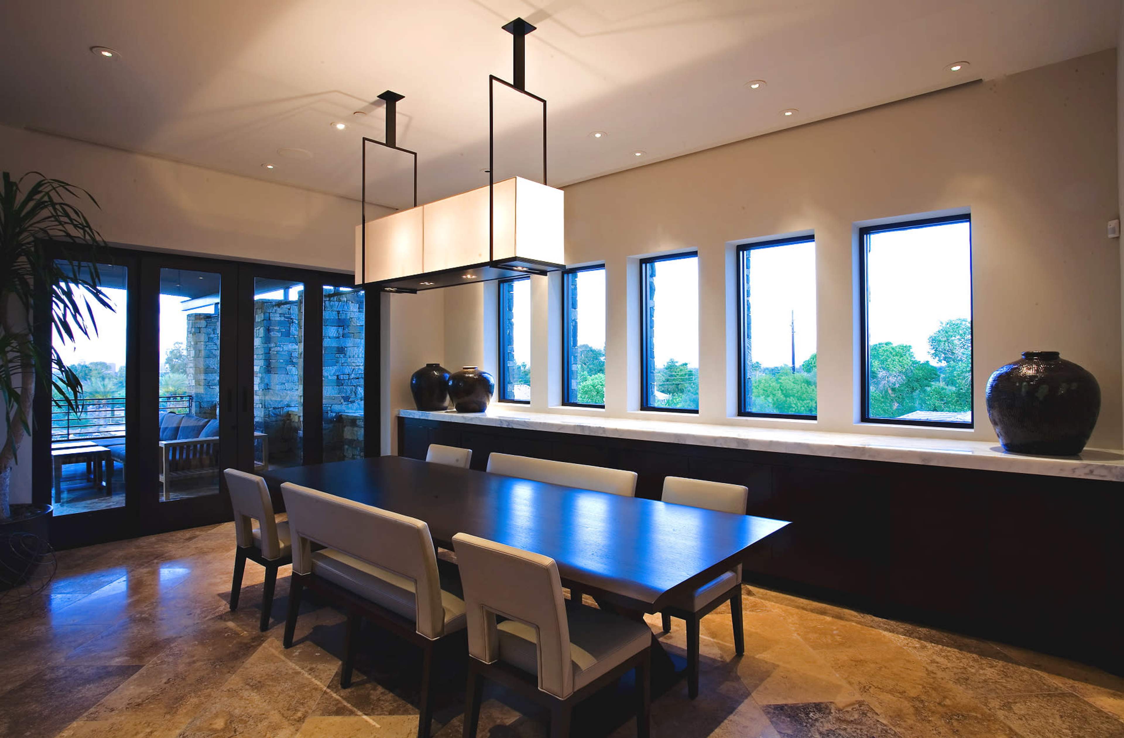 A spacious dining room features a long wooden table surrounded by upholstered chairs, with large windows allowing natural light and views of greenery.