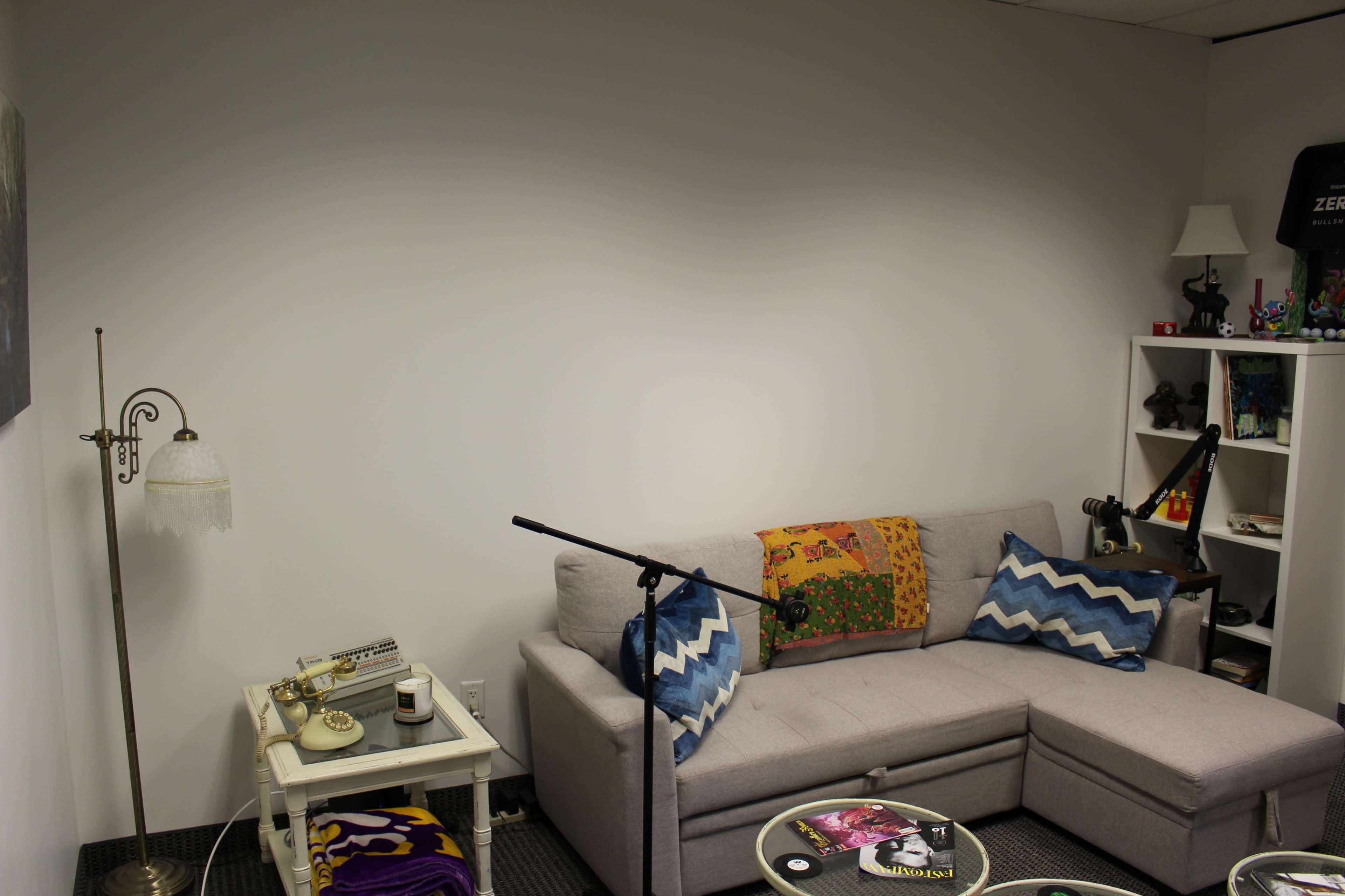 The image shows a small, sparsely furnished room featuring a gray sectional sofa with decorative pillows, a microphone on a stand, a lamp, and a white shelf filled with various items.
