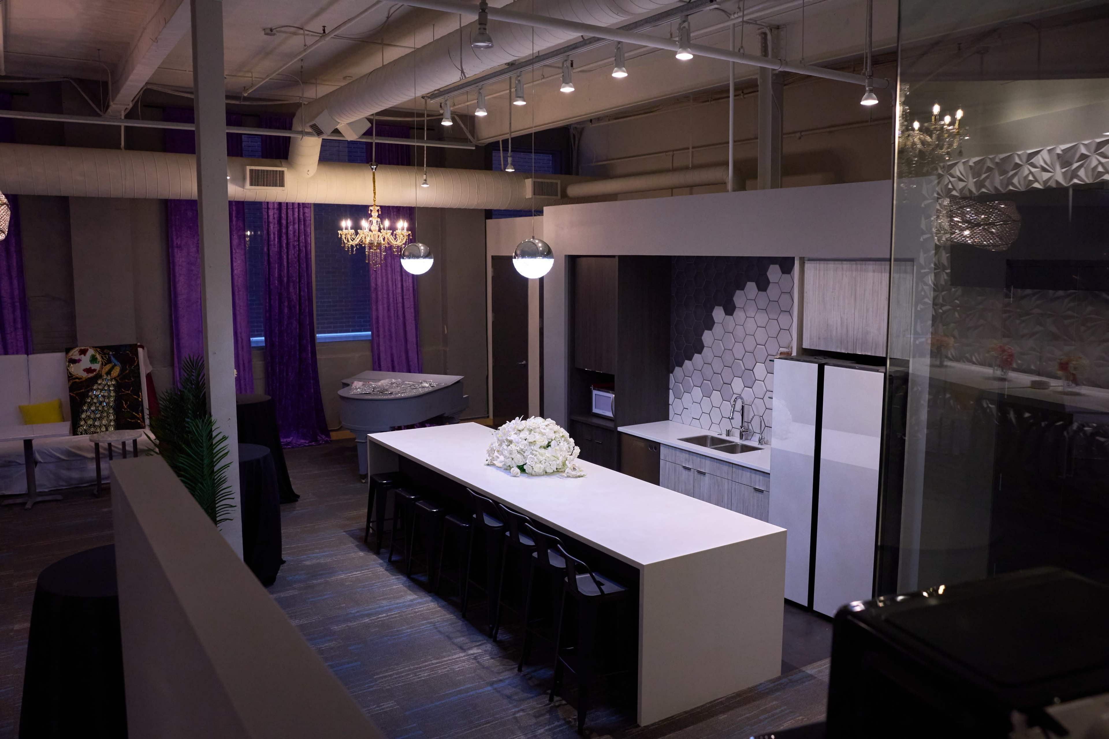 The image shows a modern kitchen and dining area with a large white island, black bar stools, and purple curtains.