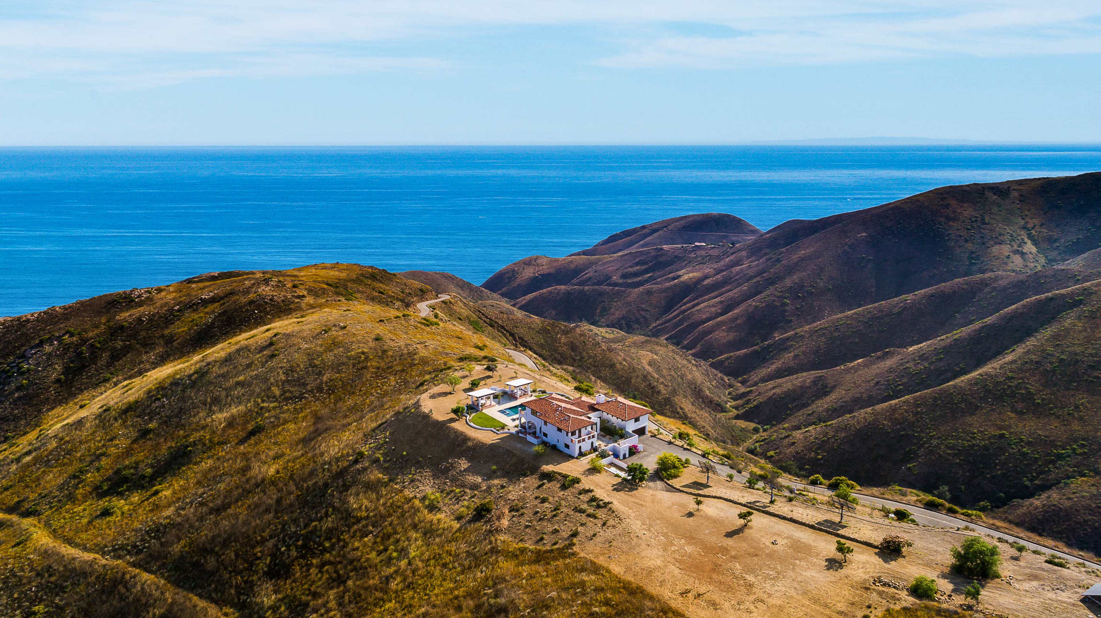 A large house sits on a hillside overlooking the ocean, surrounded by rolling brown hills.