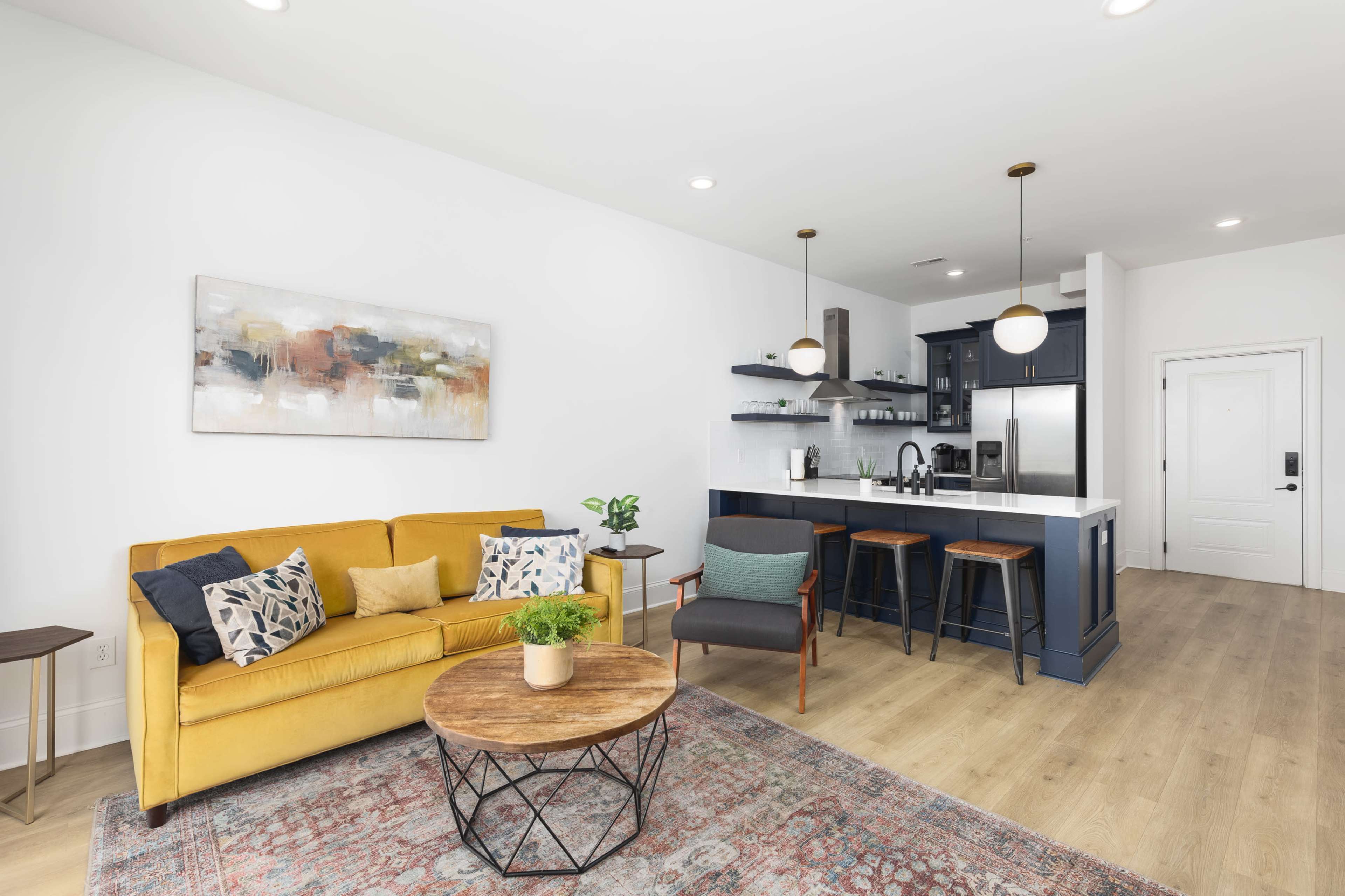 A modern living area features a mustard yellow sofa, a round wooden coffee table, and a kitchen with dark cabinetry and bar seating.