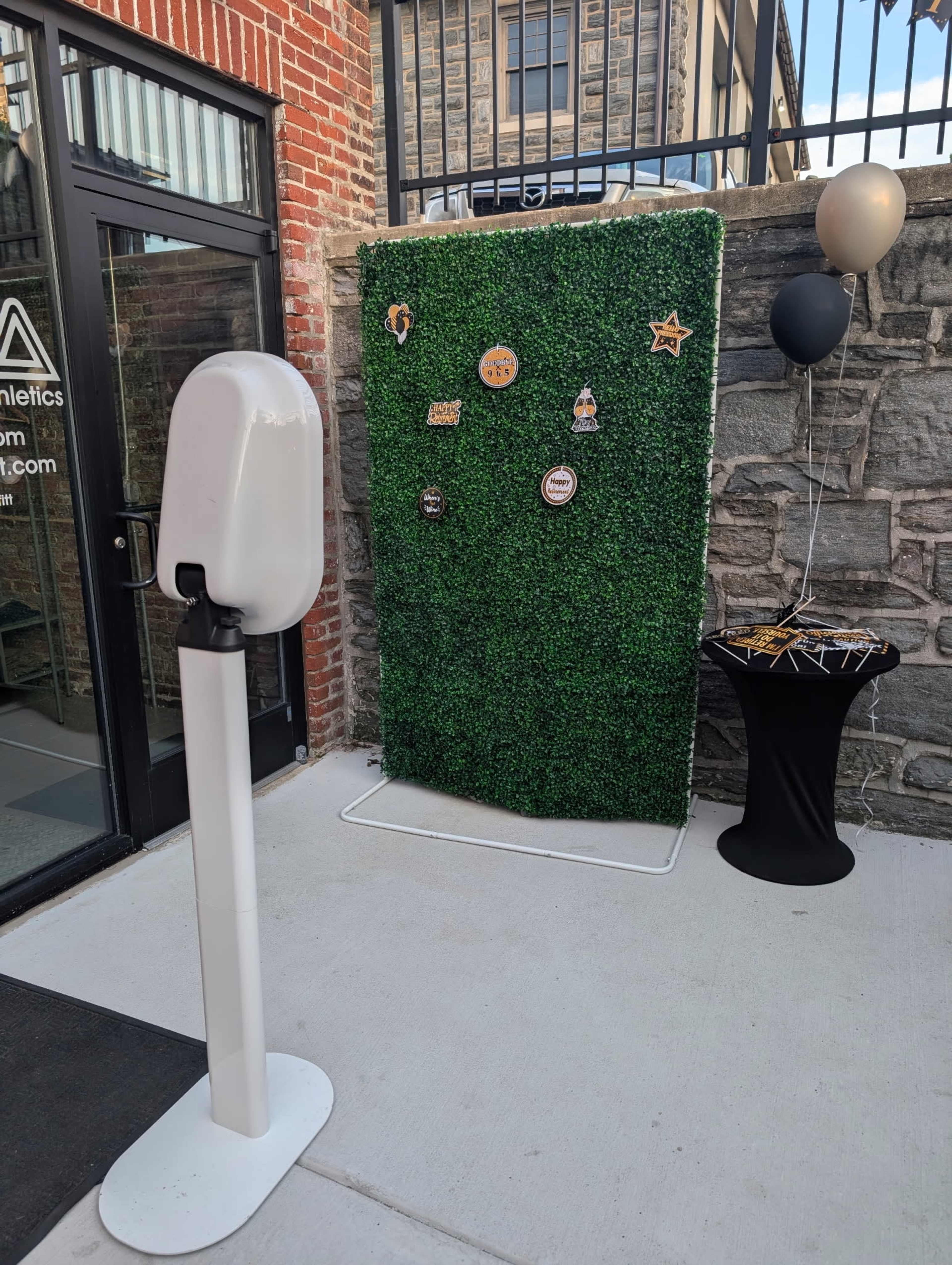 The image shows a green artificial hedge backdrop with various decorative elements, placed outside a building near a hand sanitizer dispenser and a table with a black tablecloth.
