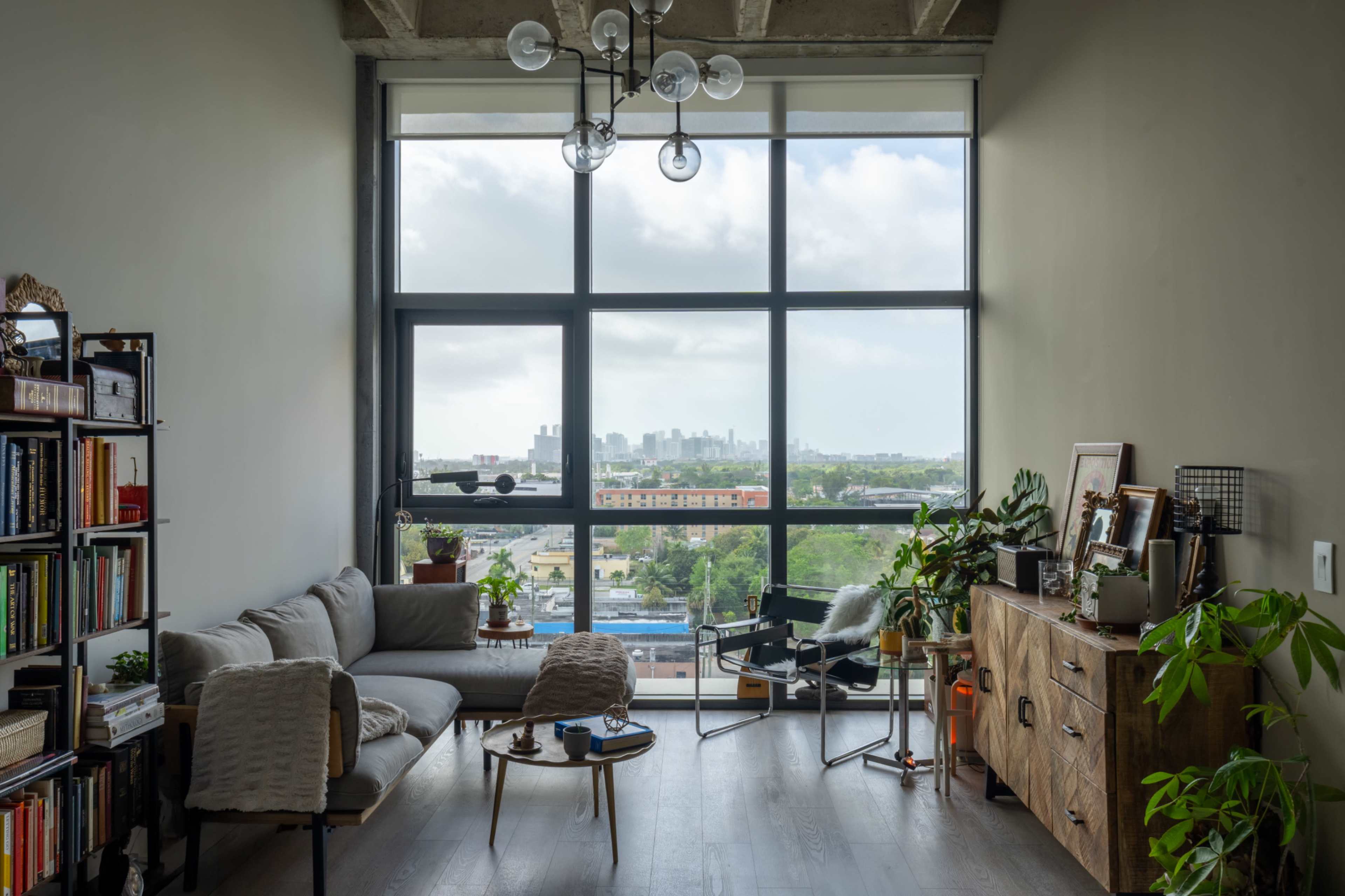A modern living room features a large window with a city view, a gray sofa, a wooden sideboard, and indoor plants.